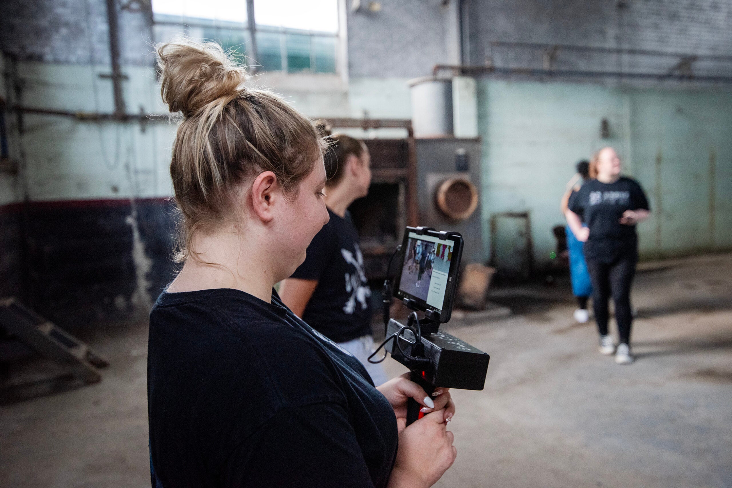 Person holding a camera rig with a smartphone filming two people standing in a large, industrial indoor space with concrete floors and worn walls.