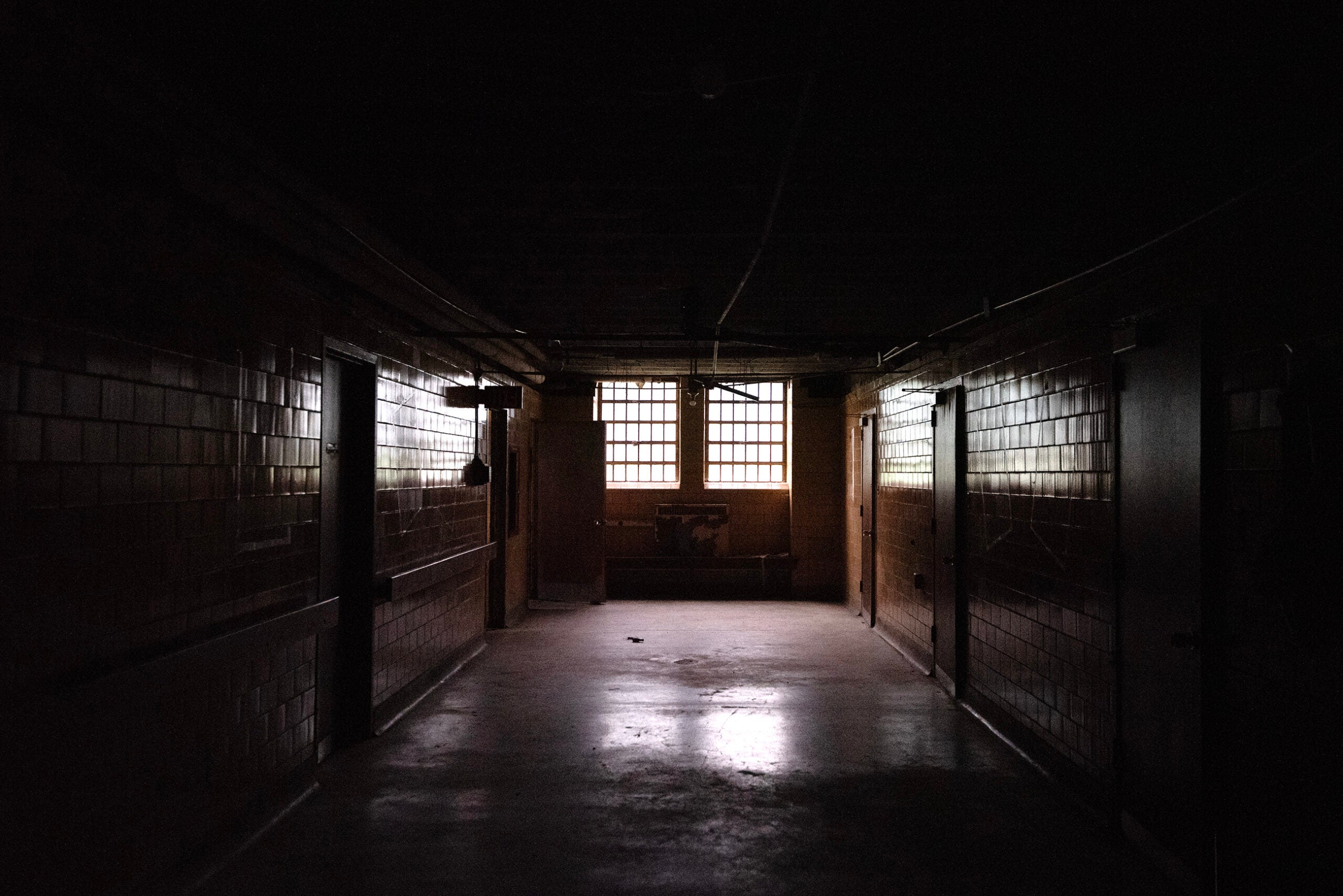 Dimly lit hallway with tiled walls, multiple doors on each side, and barred windows at the far end letting in limited natural light.