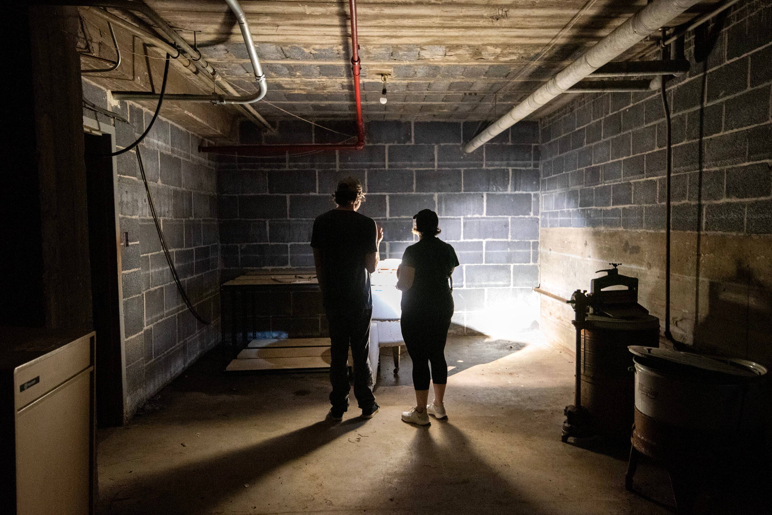 Two people stand in a dimly lit, unfinished basement or utility room with brick walls, facing a bright spot of light on a table.