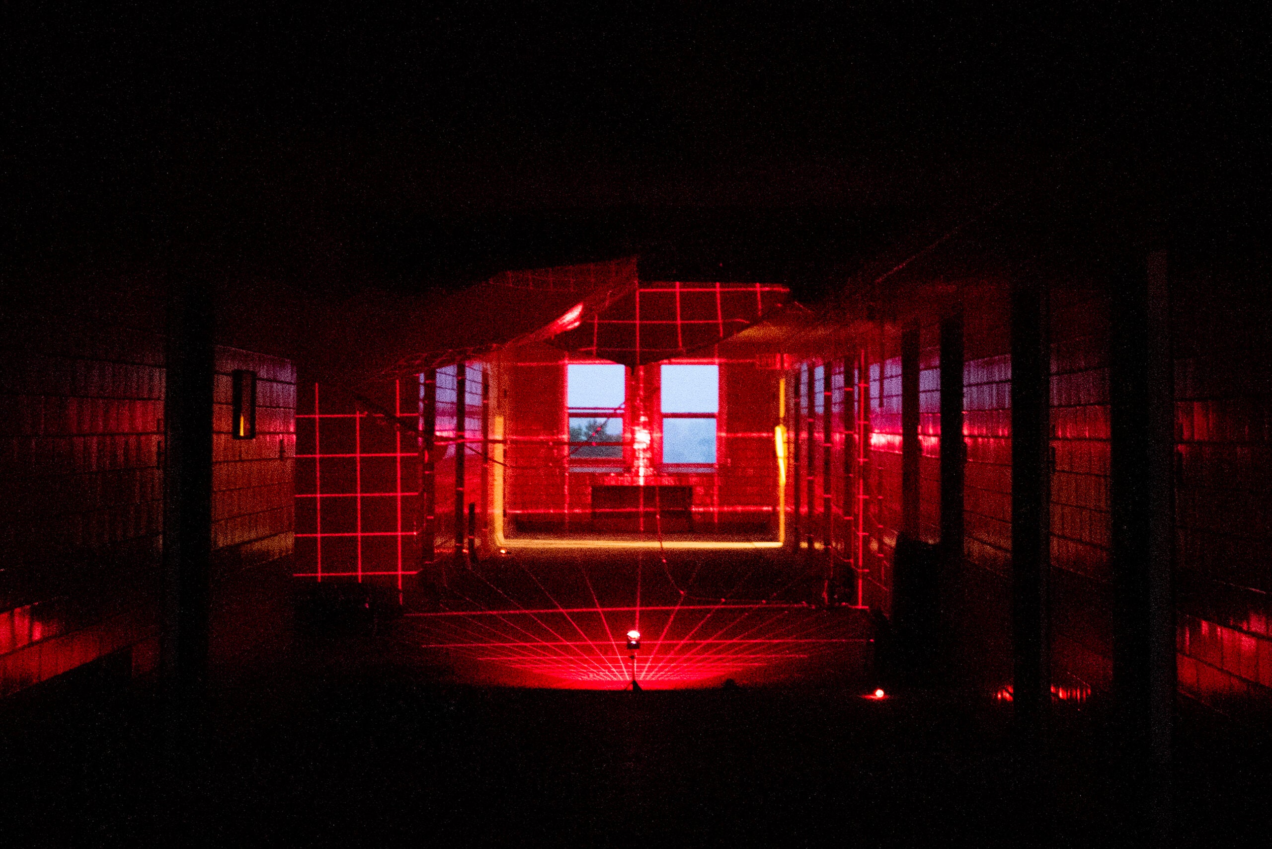 A dark hallway illuminated by a red laser grid pattern, with the grid lines projecting onto the walls, ceiling, and floor; windows are visible at the end of the hallway.