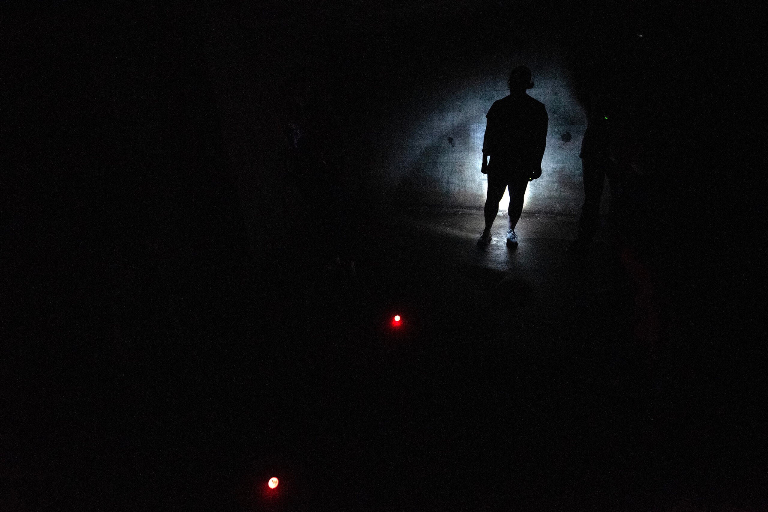A person stands in partial darkness illuminated by a flashlight, with two small red lights visible on the ground nearby.