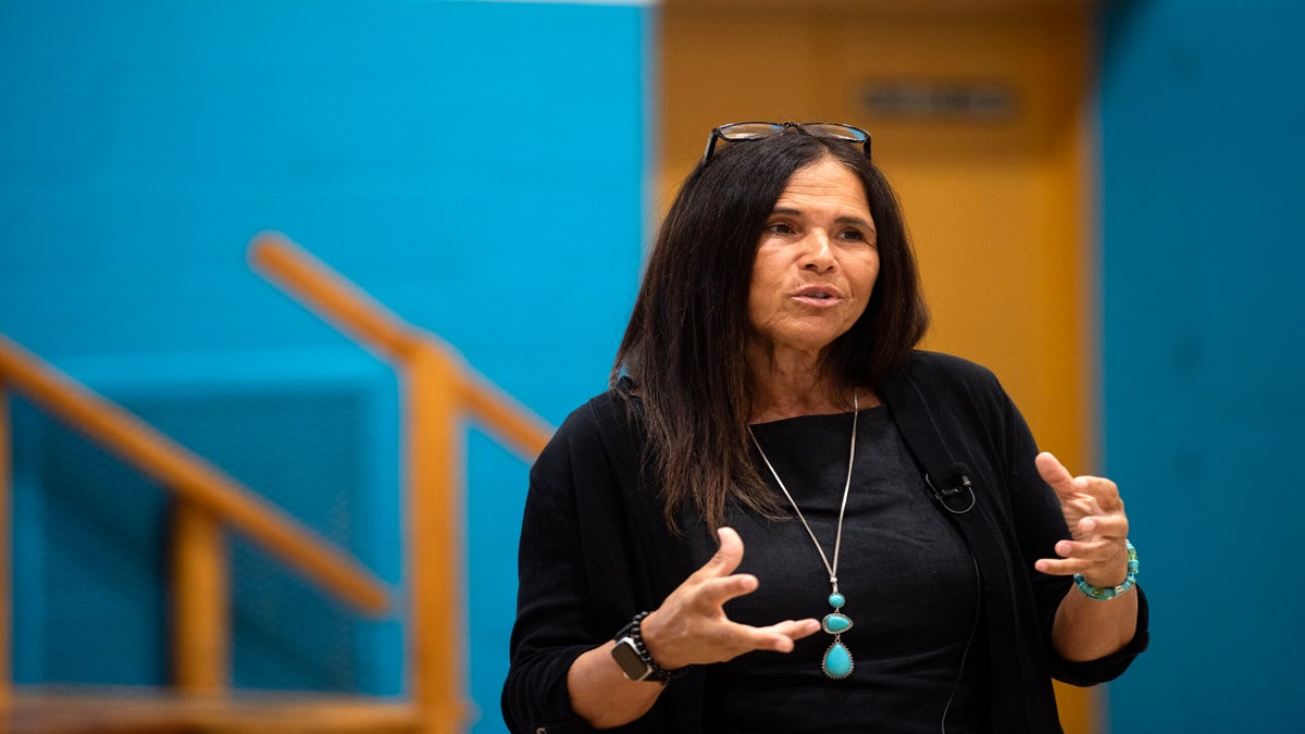 A woman with long dark hair, glasses on her head, and a turquoise necklace speaks, gesturing with her hands, in an indoor setting with blue and yellow walls.