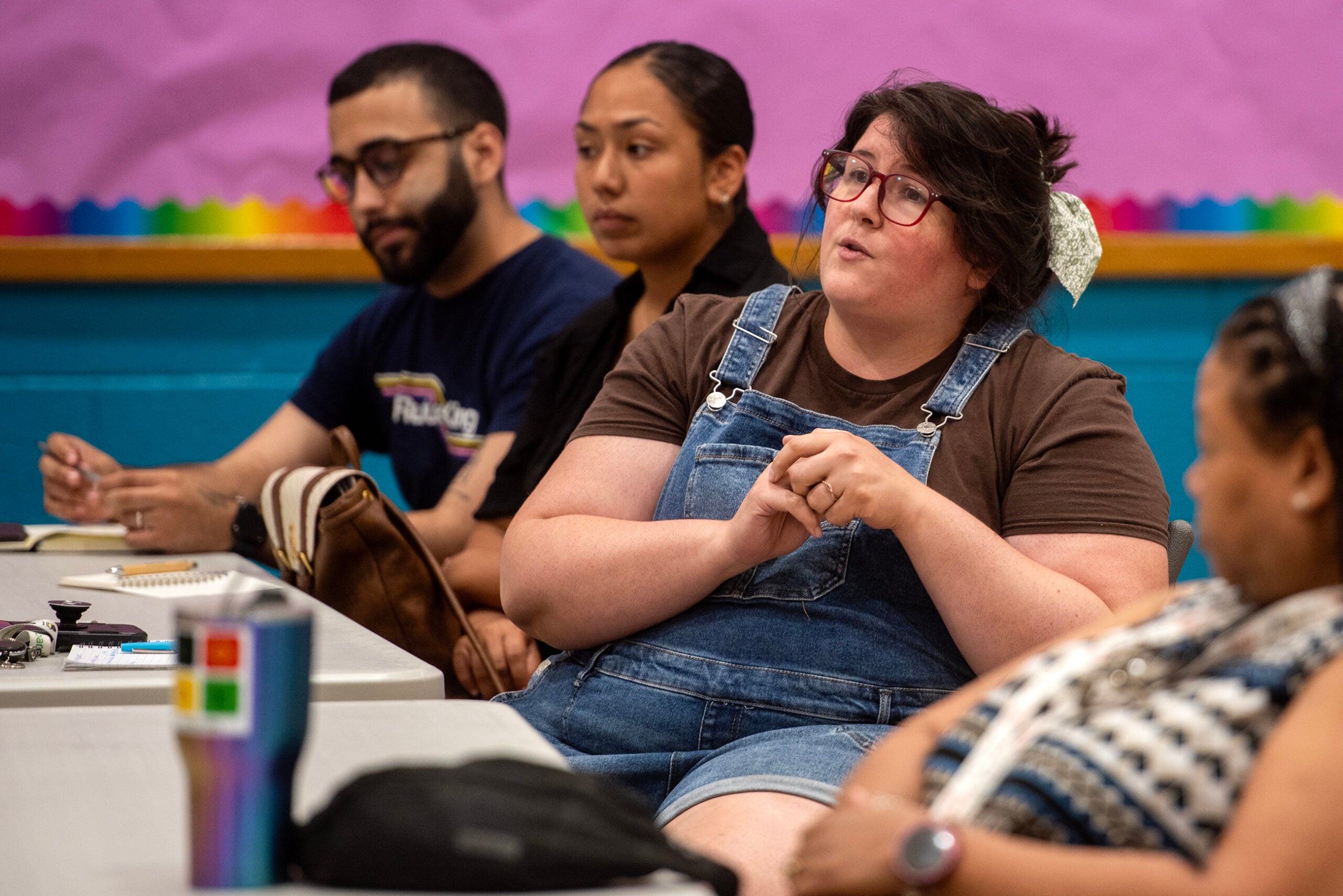 A group of people sit at tables in a classroom; one person in denim overalls speaks while others listen and take notes.