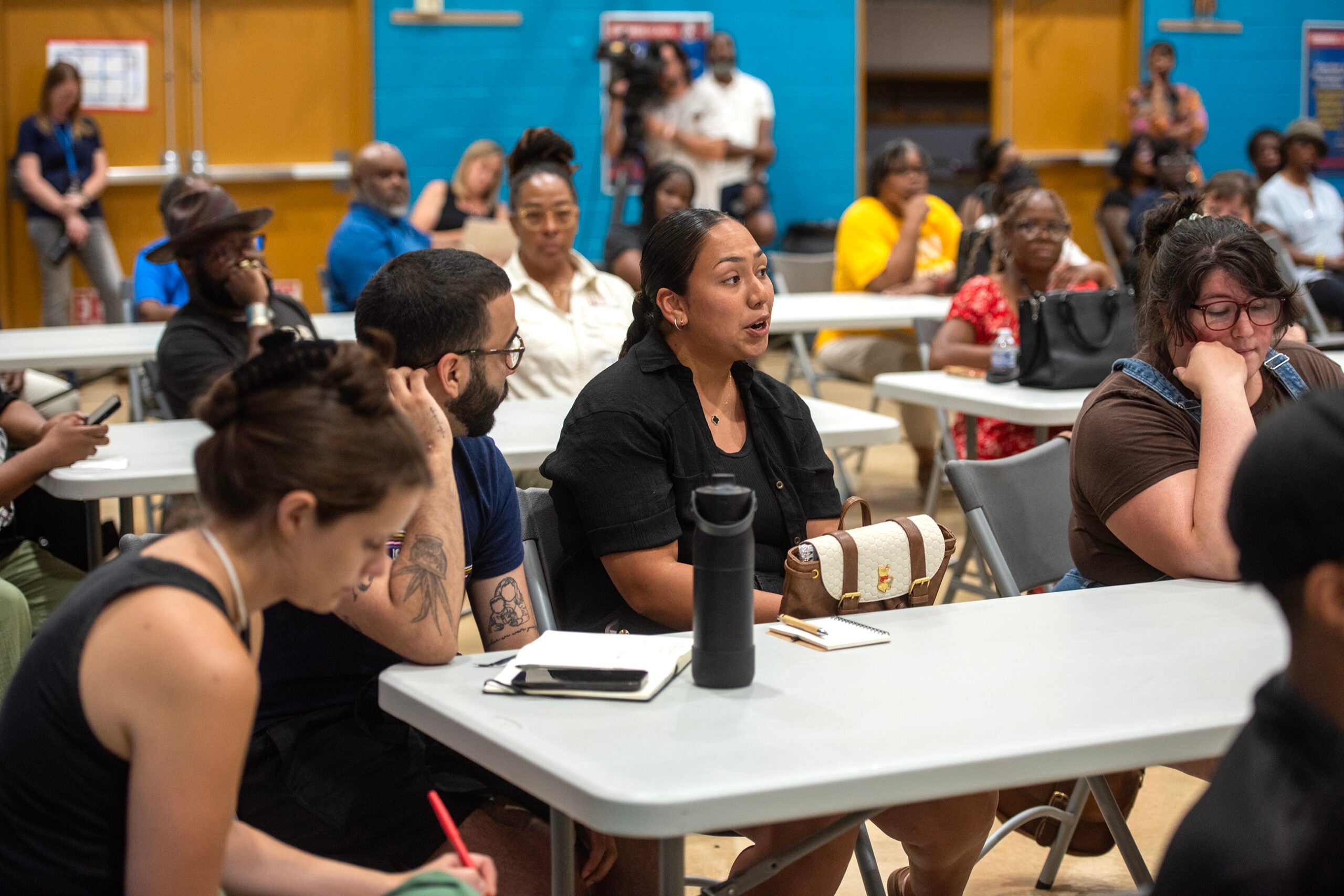 People seated at tables in a community meeting, with one woman speaking while others listen and take notes.