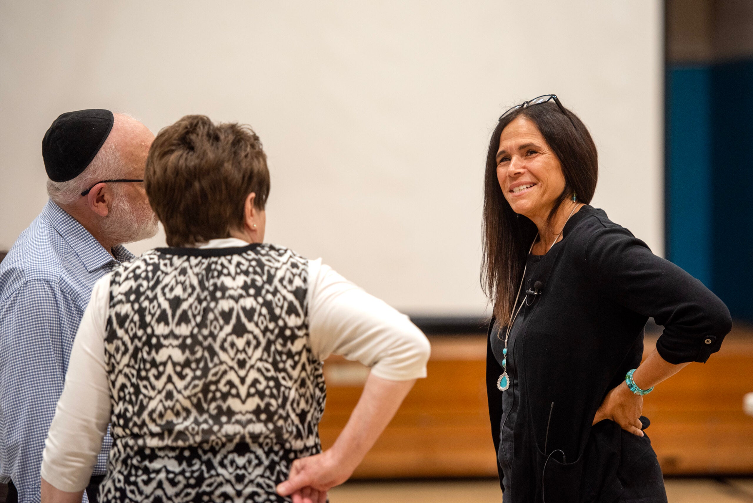 A woman smiles while talking to two people in a casual indoor setting, with a blurred background featuring a large screen and wooden benches.