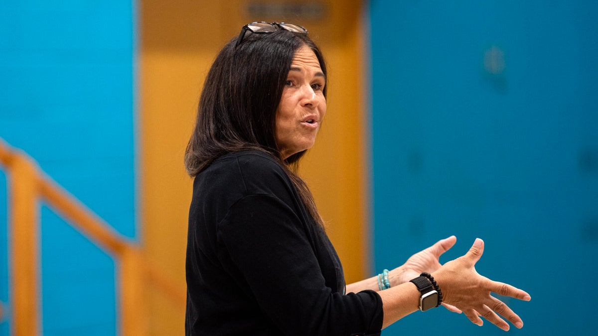 A woman with long dark hair speaks while gesturing with her hands indoors; she wears a black top, watch, and glasses on her head. Blue and yellow walls are visible in the background.