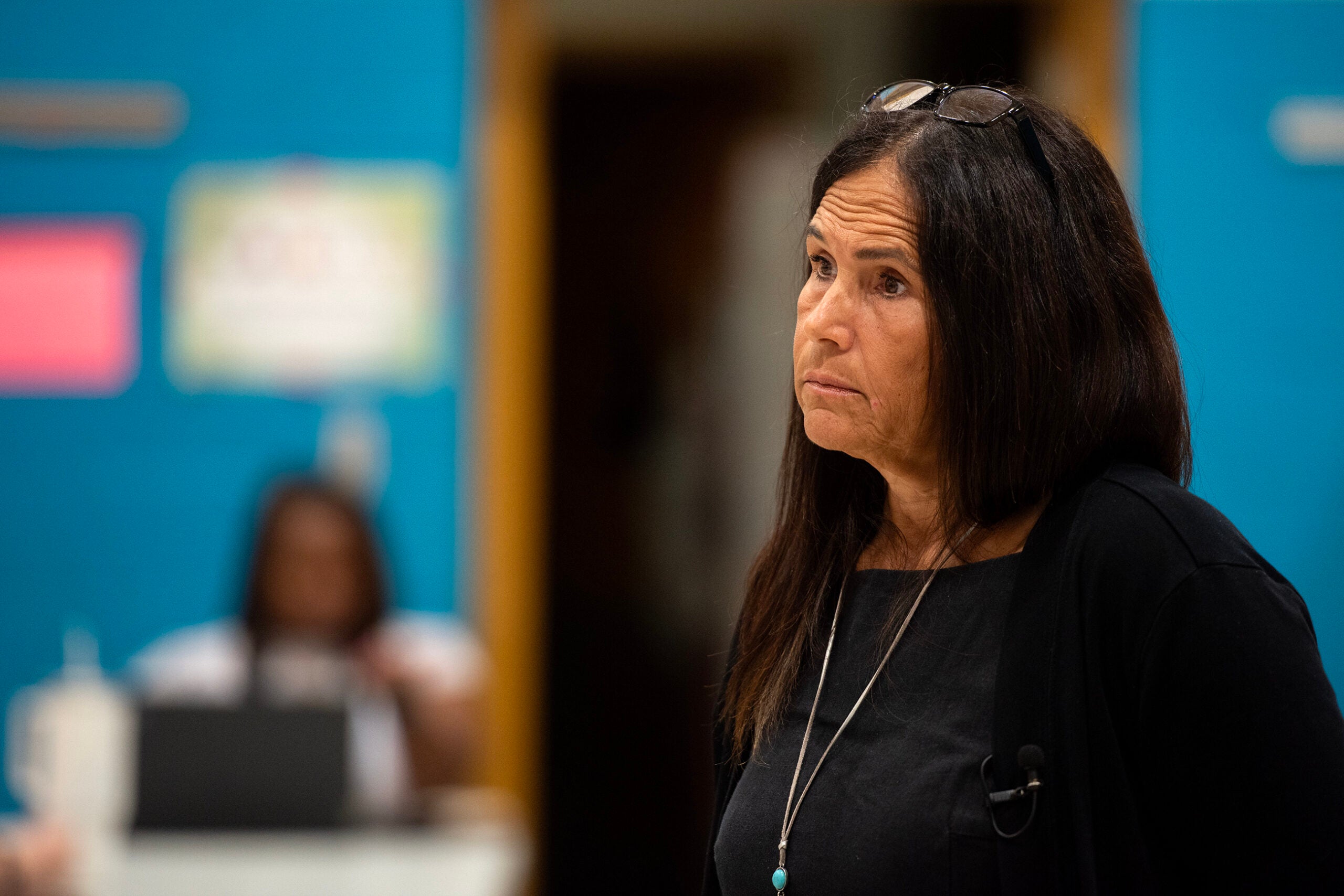 A woman with long dark hair and glasses on her head stands indoors, looking to the side; a blurred person works at a desk in the background.