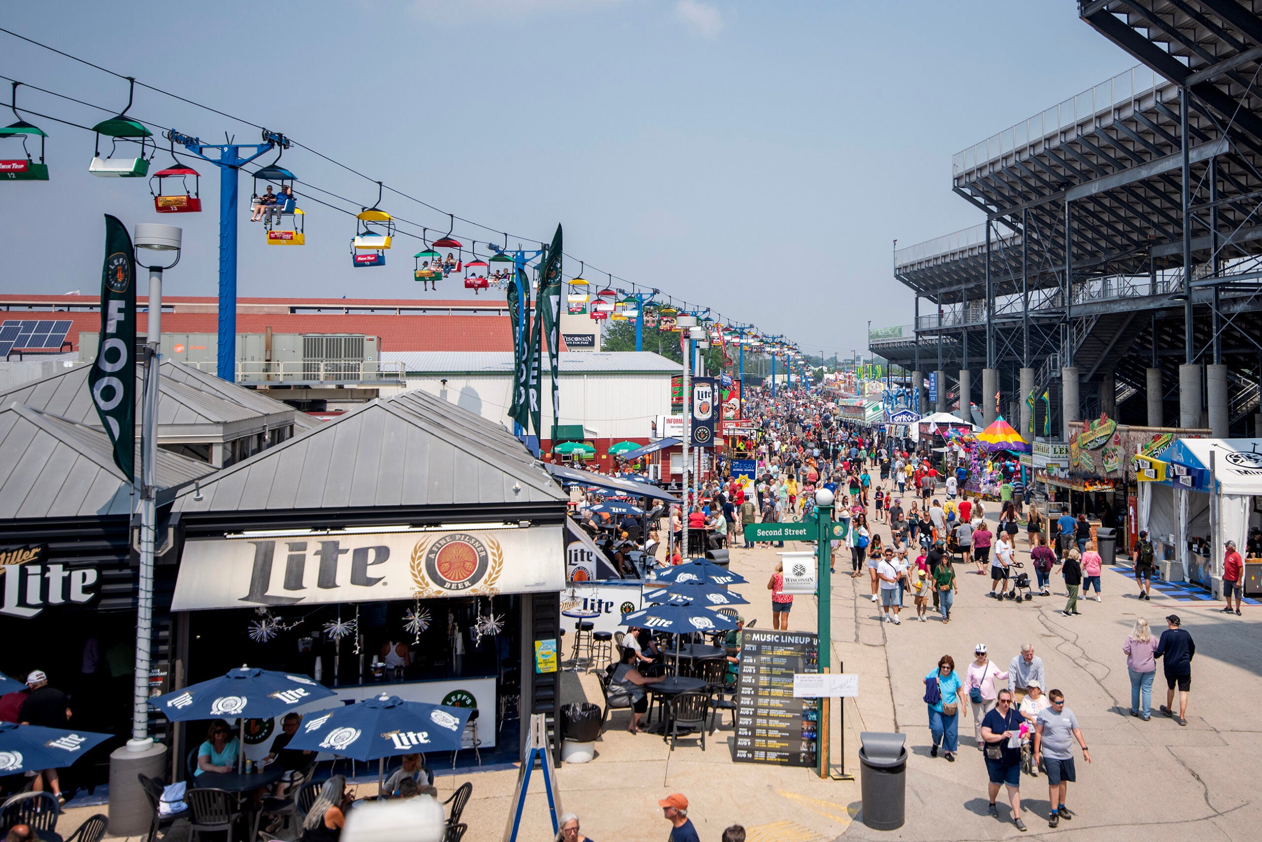 A crowded outdoor fairground with food stands, people walking, a large stadium on the right, and colorful gondolas overhead.