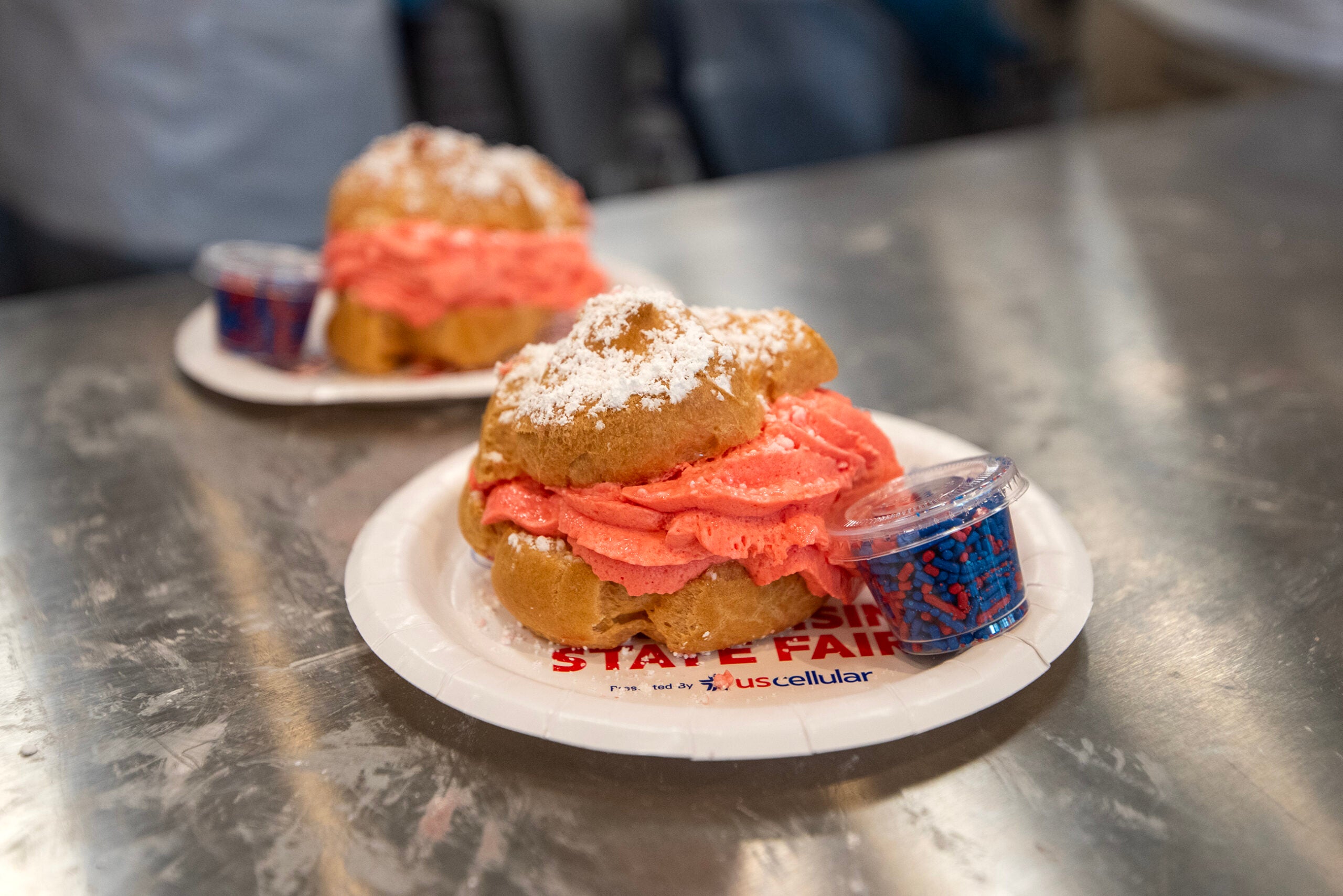 Two cream puffs with pink filling and powdered sugar are served on paper plates with a small container of red and blue sprinkles on a metal surface.