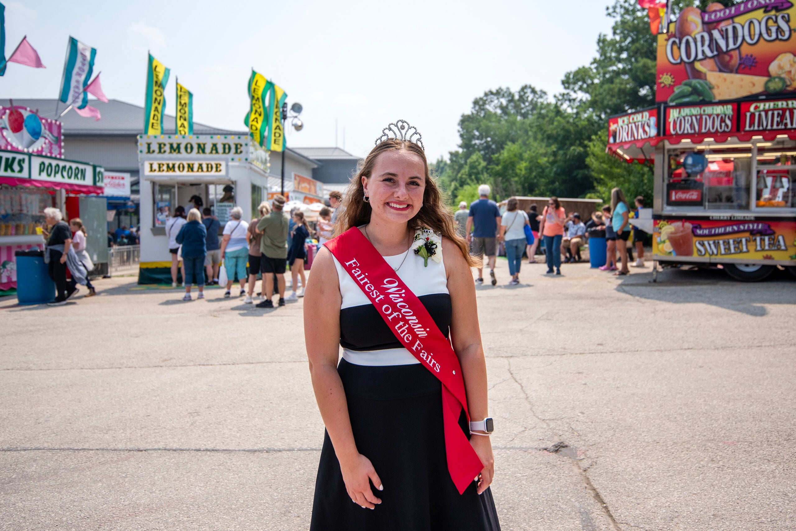A young woman wearing a crown and sash stands at a fairground, with food stalls and people in the background.