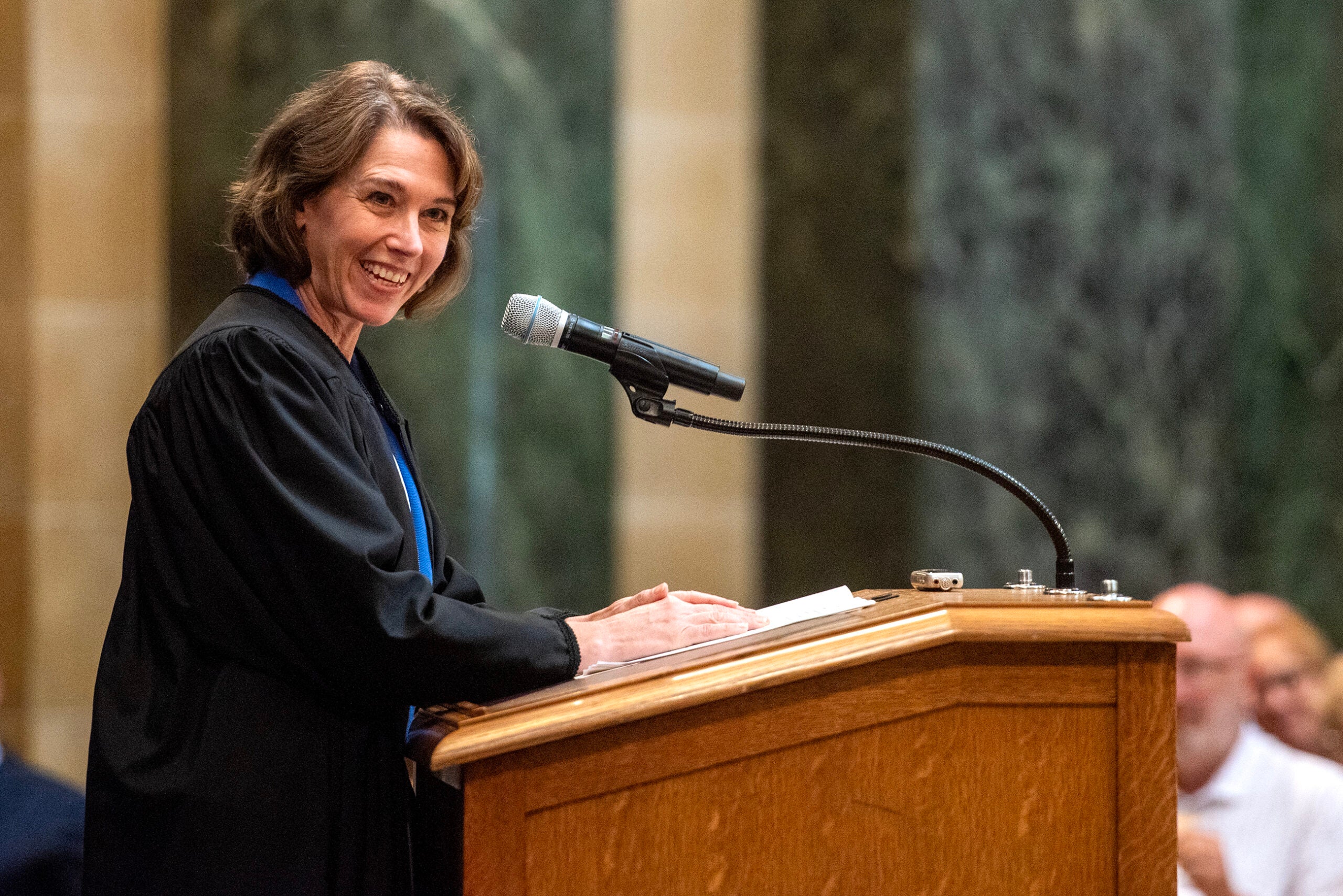 A woman in a judge’s robe speaks at a podium with a microphone in a formal setting, smiling and addressing an audience.