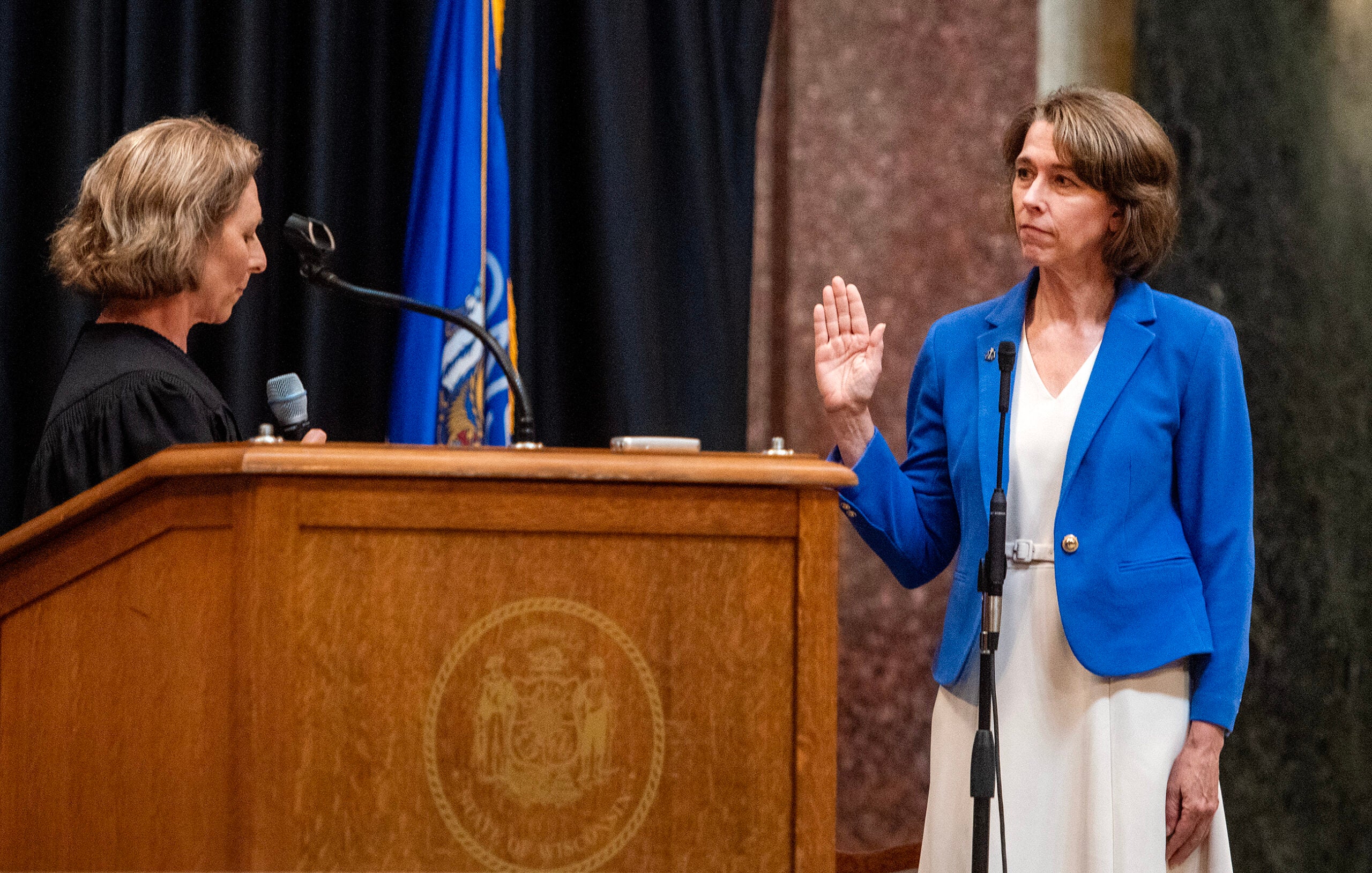 A woman in a blue blazer raises her right hand to be sworn in by a judge standing at a podium with a state seal and a blue flag in the background.