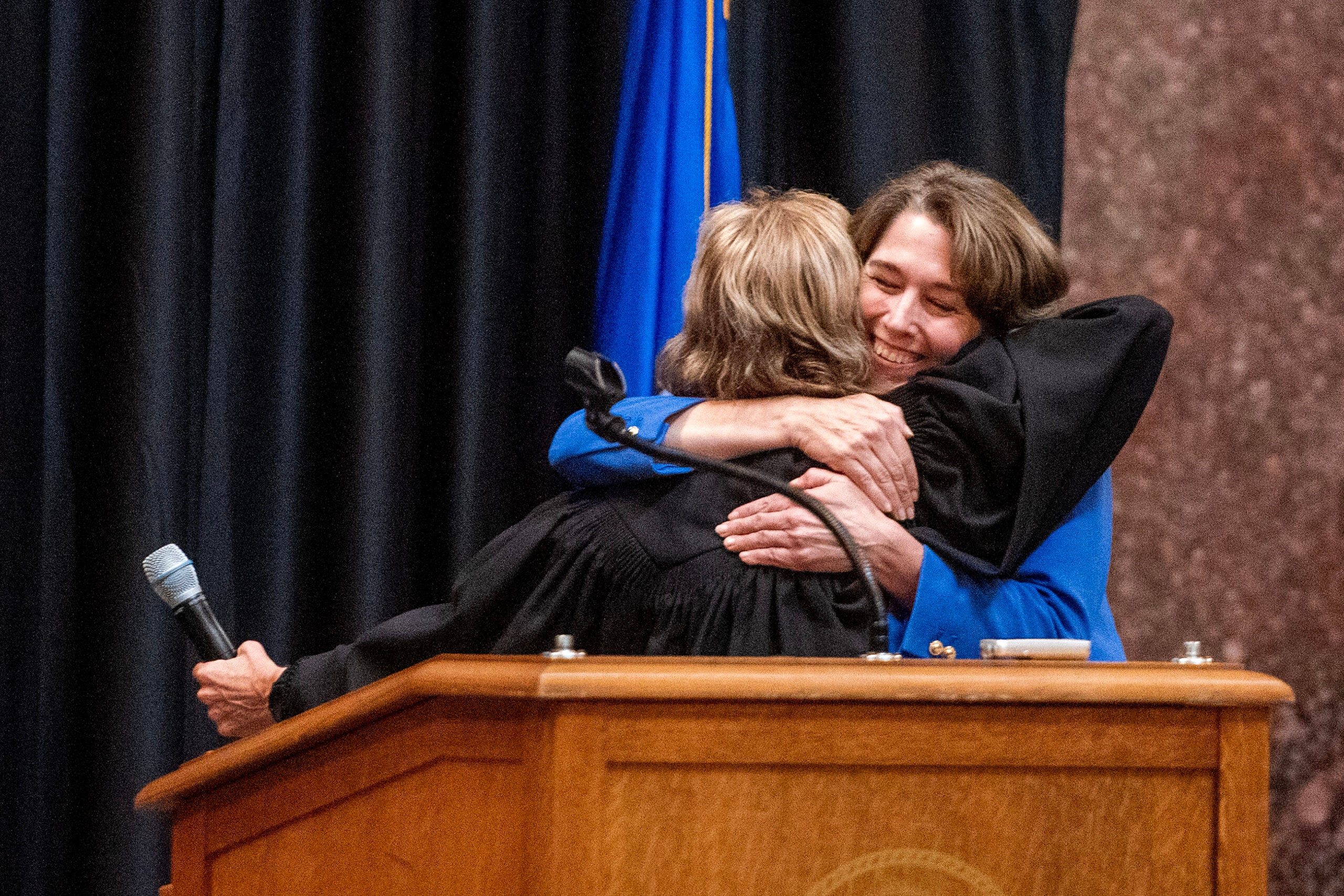 Two women embrace behind a podium; one holds a microphone, and both appear to be smiling.