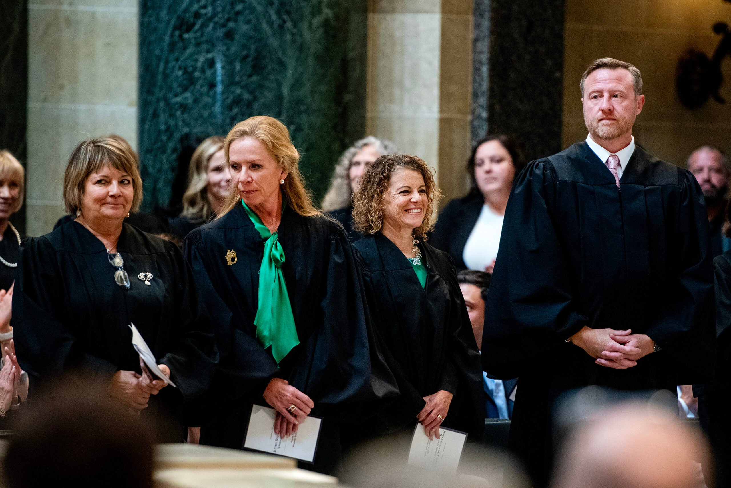 Four judges in black robes stand side by side in a formal setting, surrounded by other people, appearing attentive.