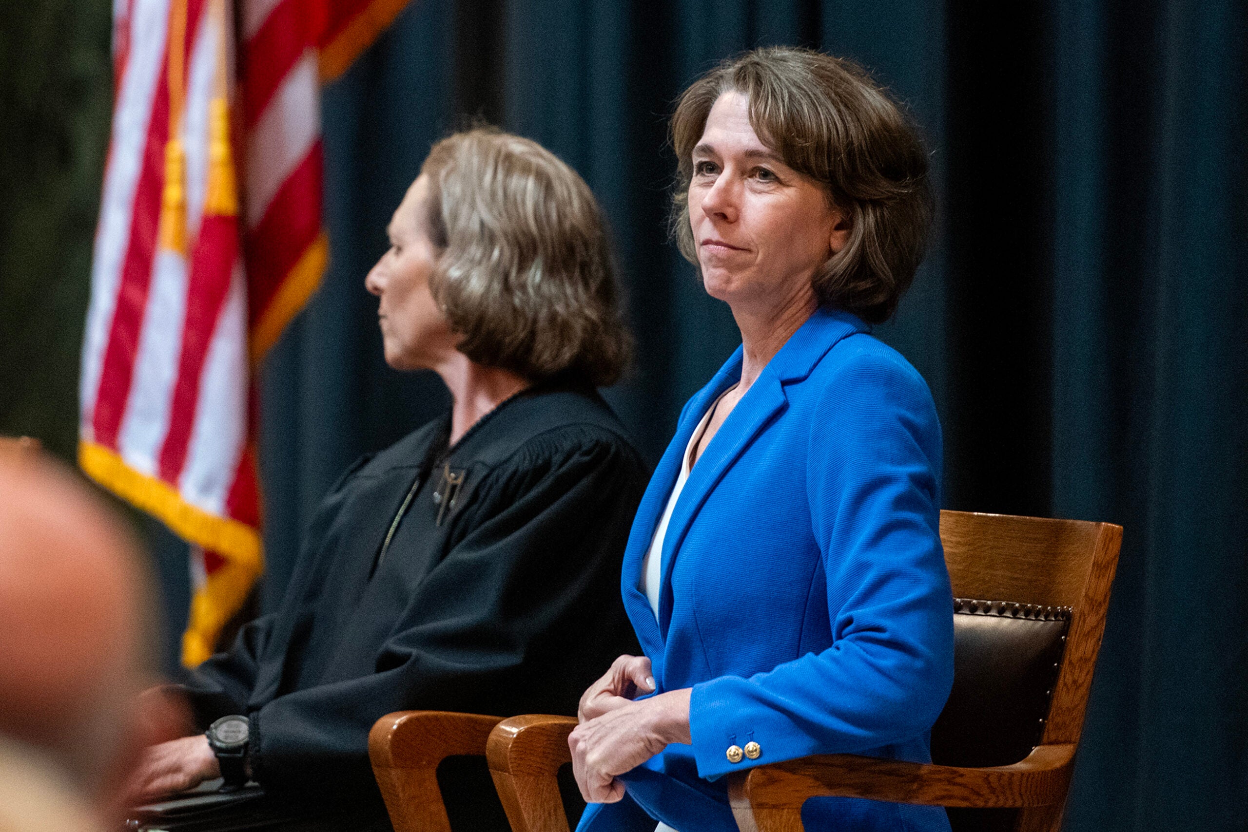 Two women sit on stage; one in a blue blazer faces forward, while the other in a black robe sits beside her facing sideways. An American flag hangs in the background.