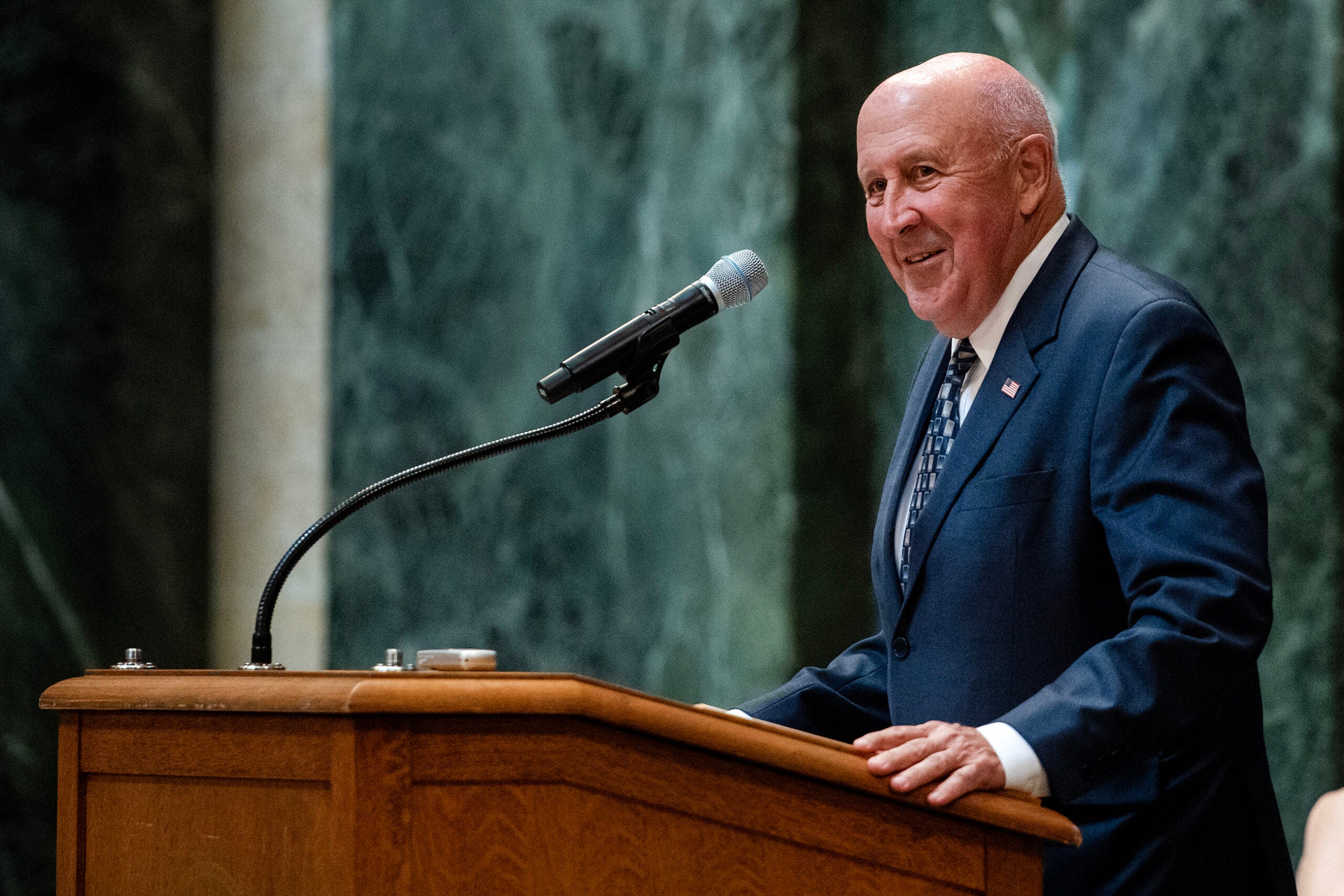 A man in a suit speaks at a podium with a microphone in a formal, marble-walled setting.