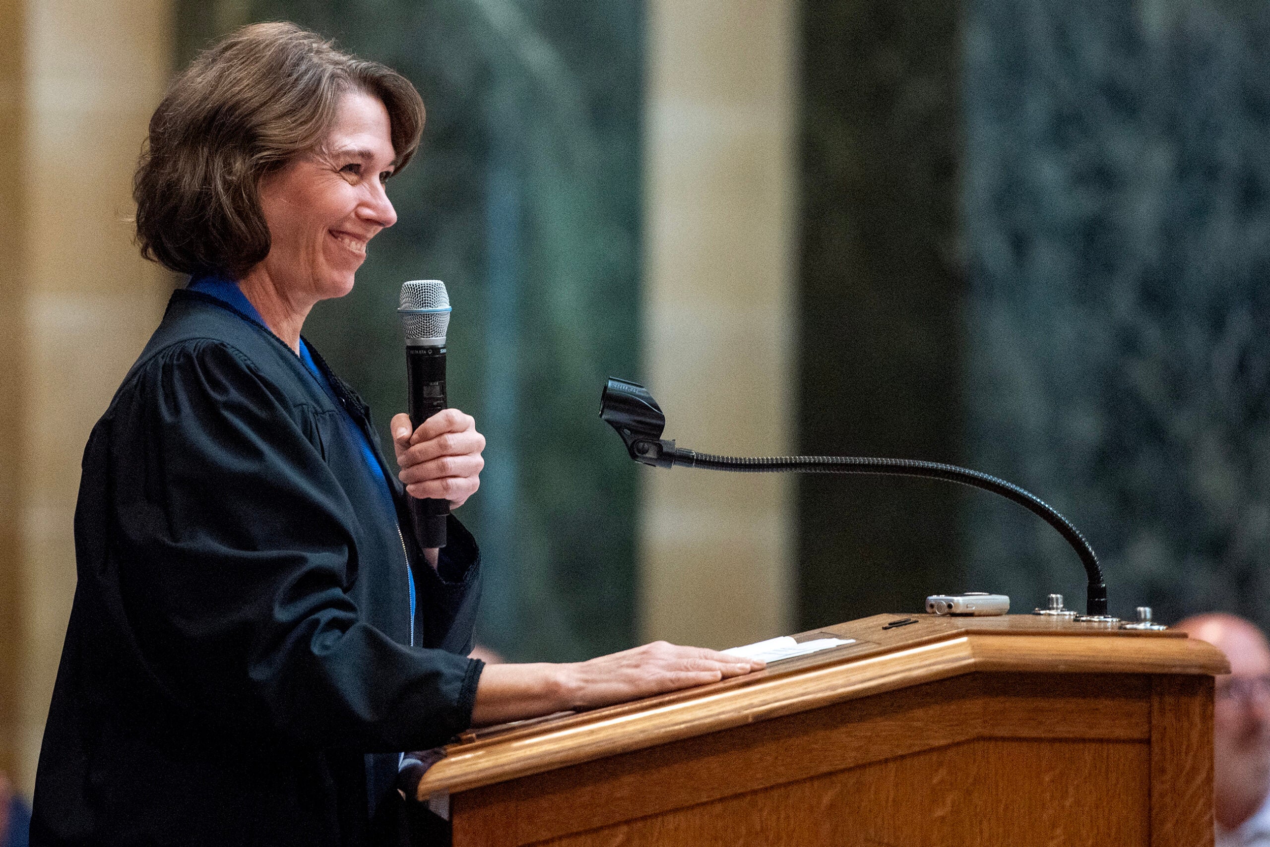 A woman in a judges robe speaks into a microphone at a wooden podium in a formal setting.