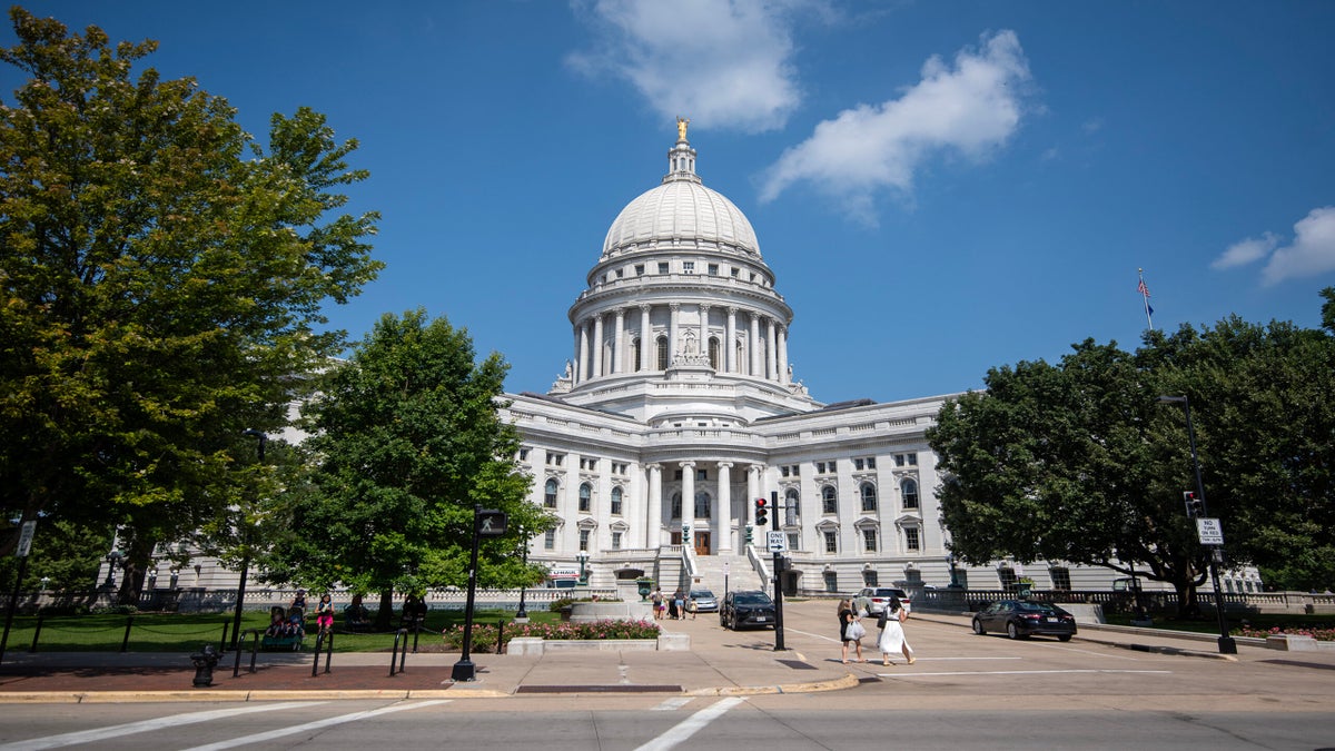 The image shows the exterior of a domed government building, with people walking near the entrance, surrounded by trees and a partly cloudy sky.