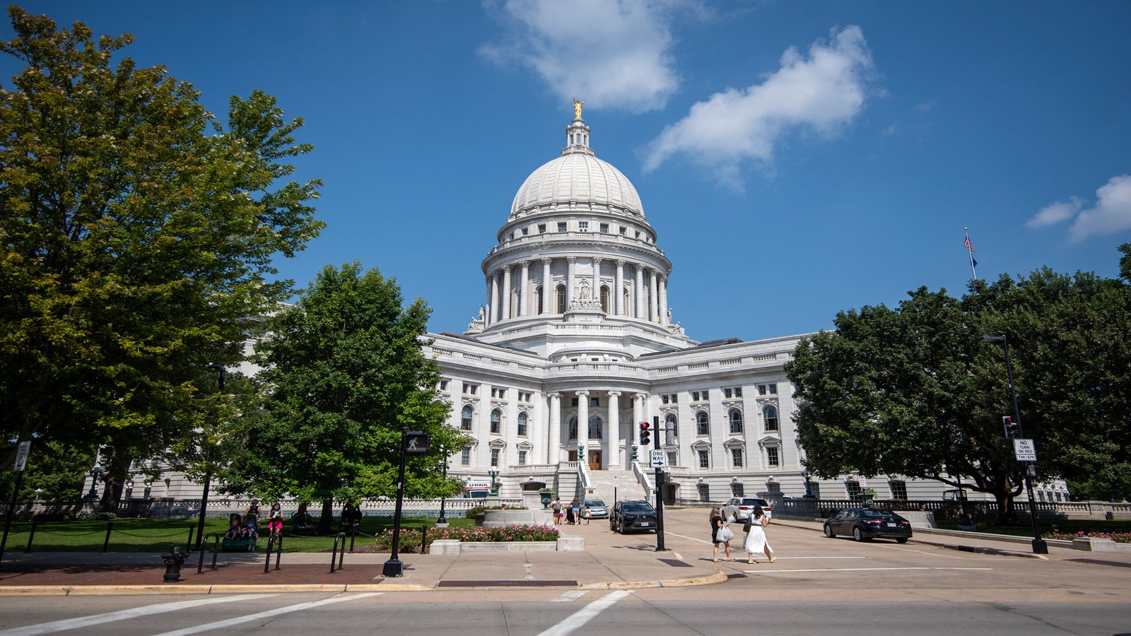 The image shows the exterior of a domed government building, with people walking near the entrance, surrounded by trees and a partly cloudy sky.