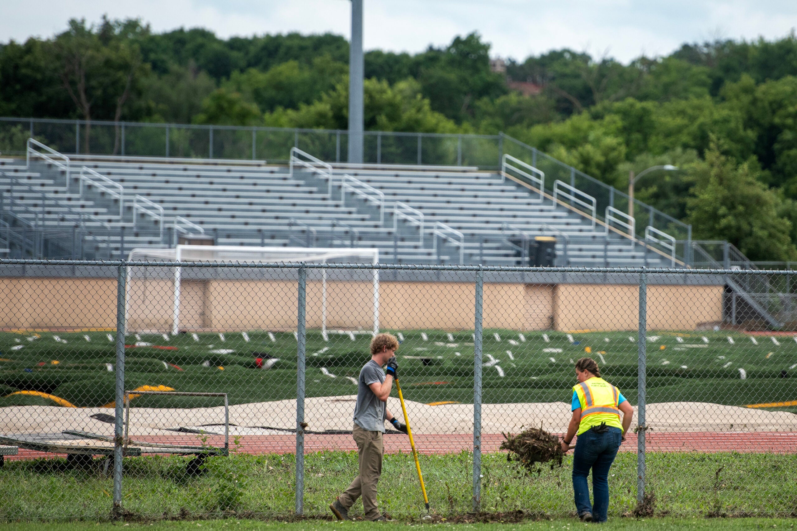 Two people work to clear grass and debris alongside a chain-link fence near an empty stadium with bleachers in the background.