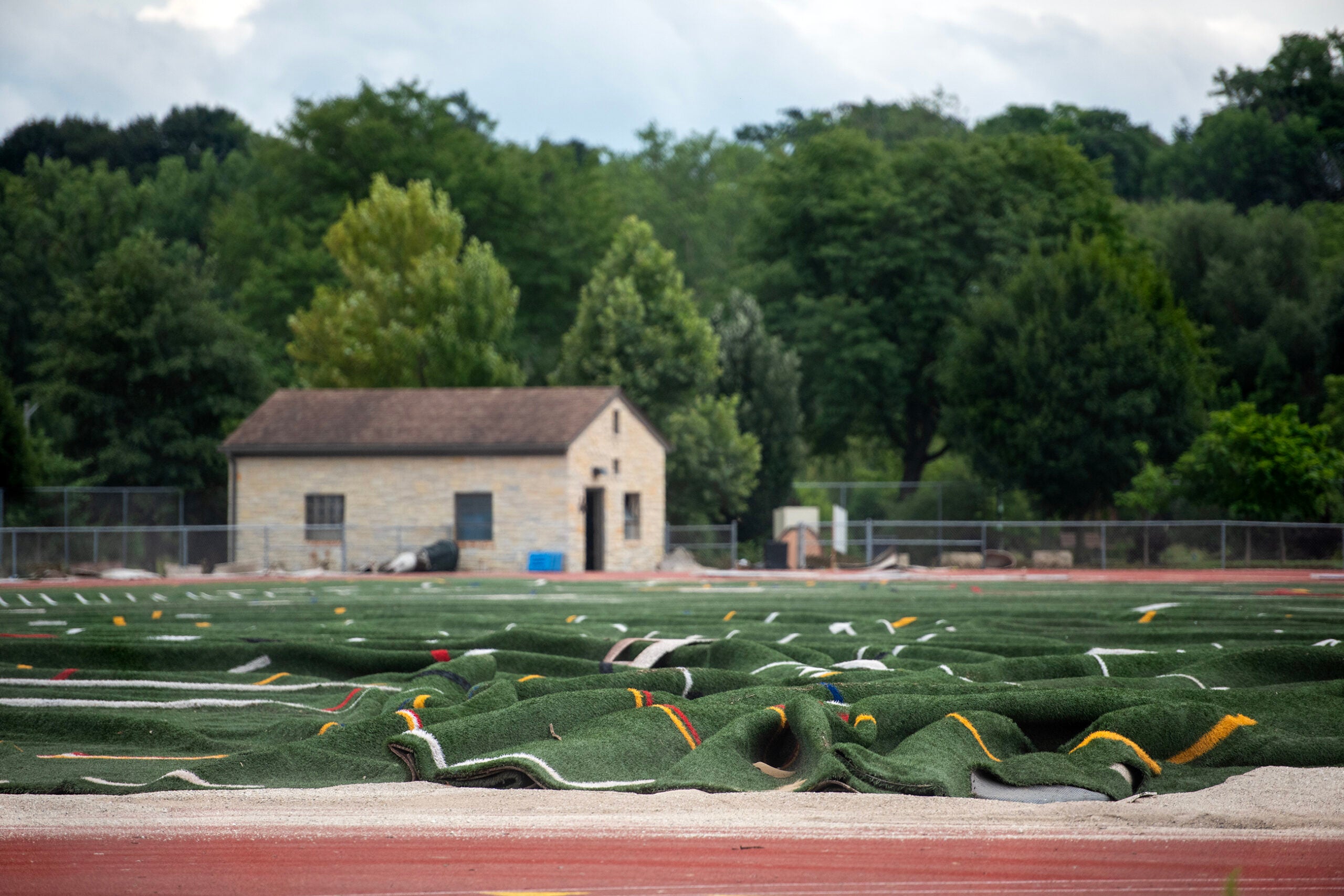 Rolled-up artificial turf lies on a sports field under renovation, with a small stone building and trees in the background.