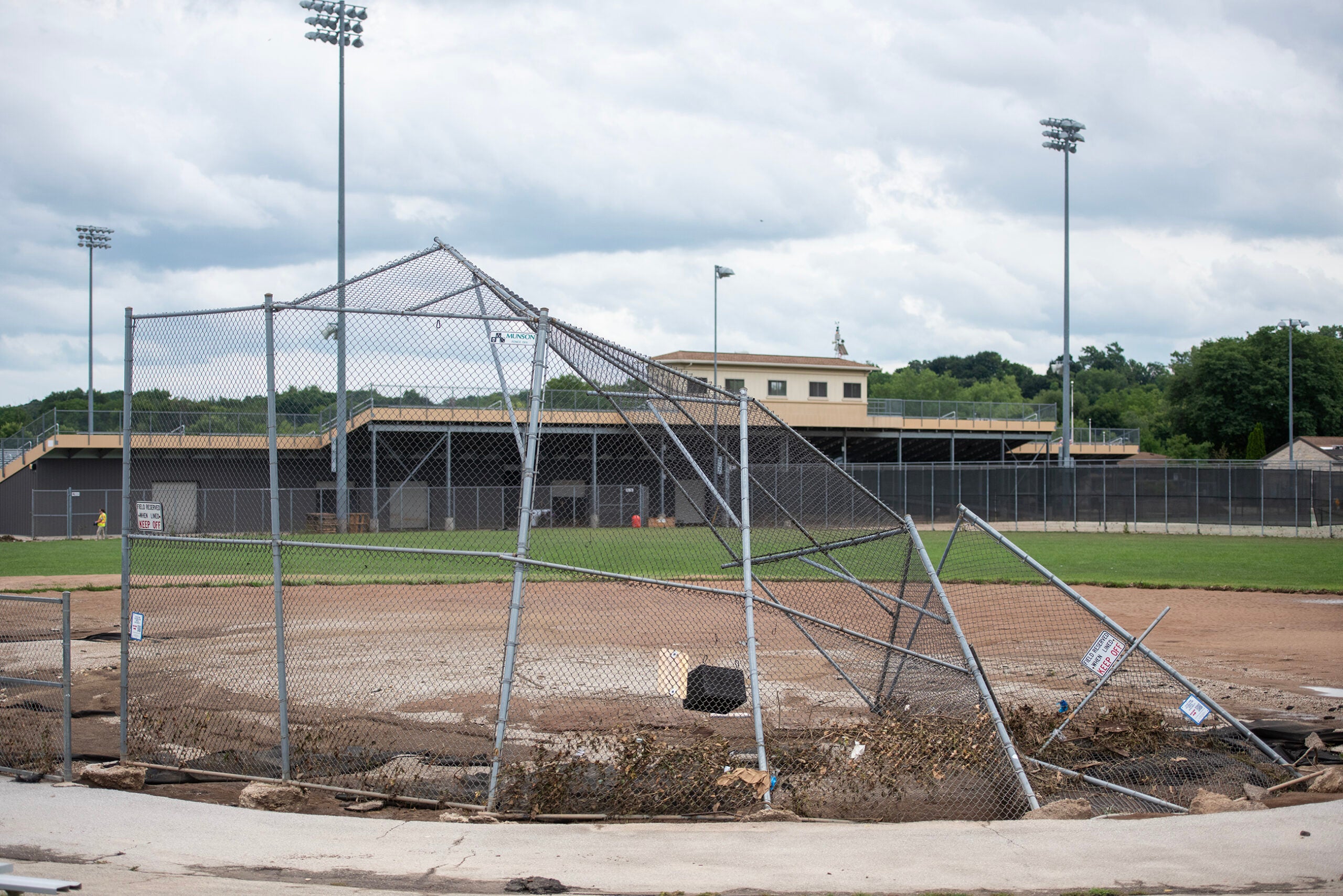 A damaged baseball field with a collapsed backstop fence, dirt exposed, and empty bleachers in the background under a cloudy sky.