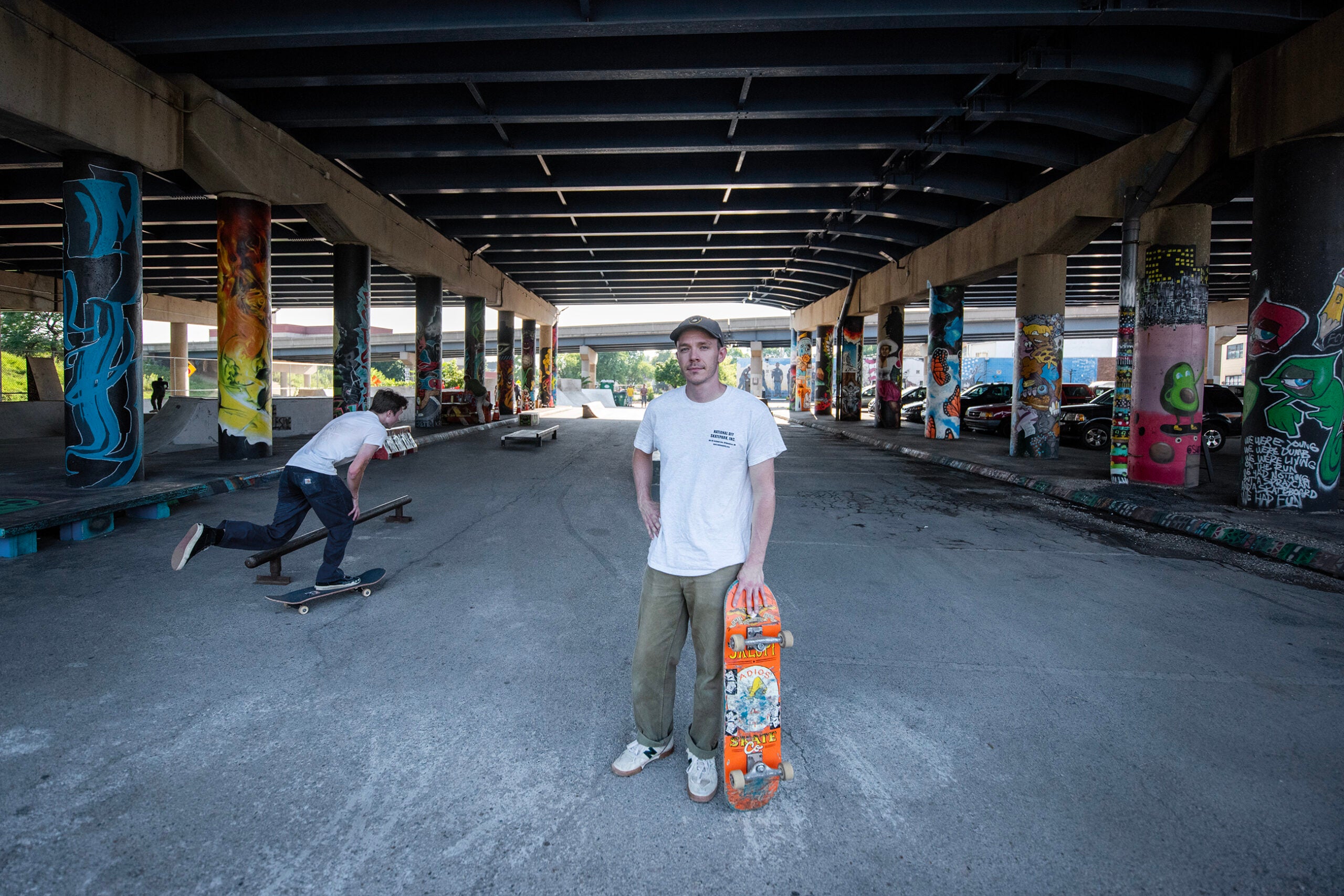 A person stands holding a skateboard under a graffiti-covered overpass while another skateboarder rides in the background.