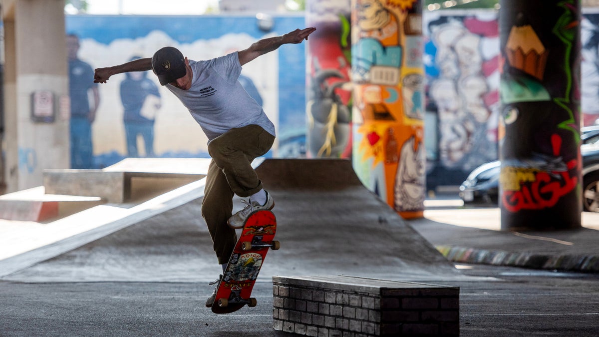 A skateboarder performs a trick over a low brick obstacle in an urban skatepark with colorful graffiti on pillars in the background.