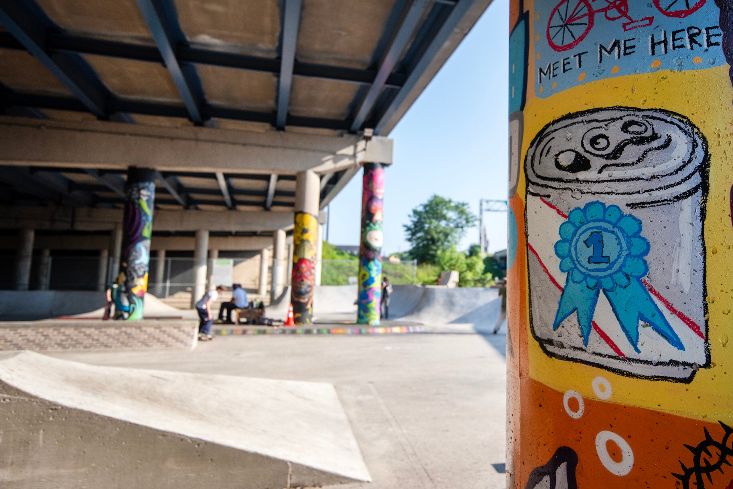 Graffiti-covered pillars under a bridge at a skatepark; foreground pillar shows a first-place medal on a can and text MEET ME HERE. Skaters and ramps visible in the background.