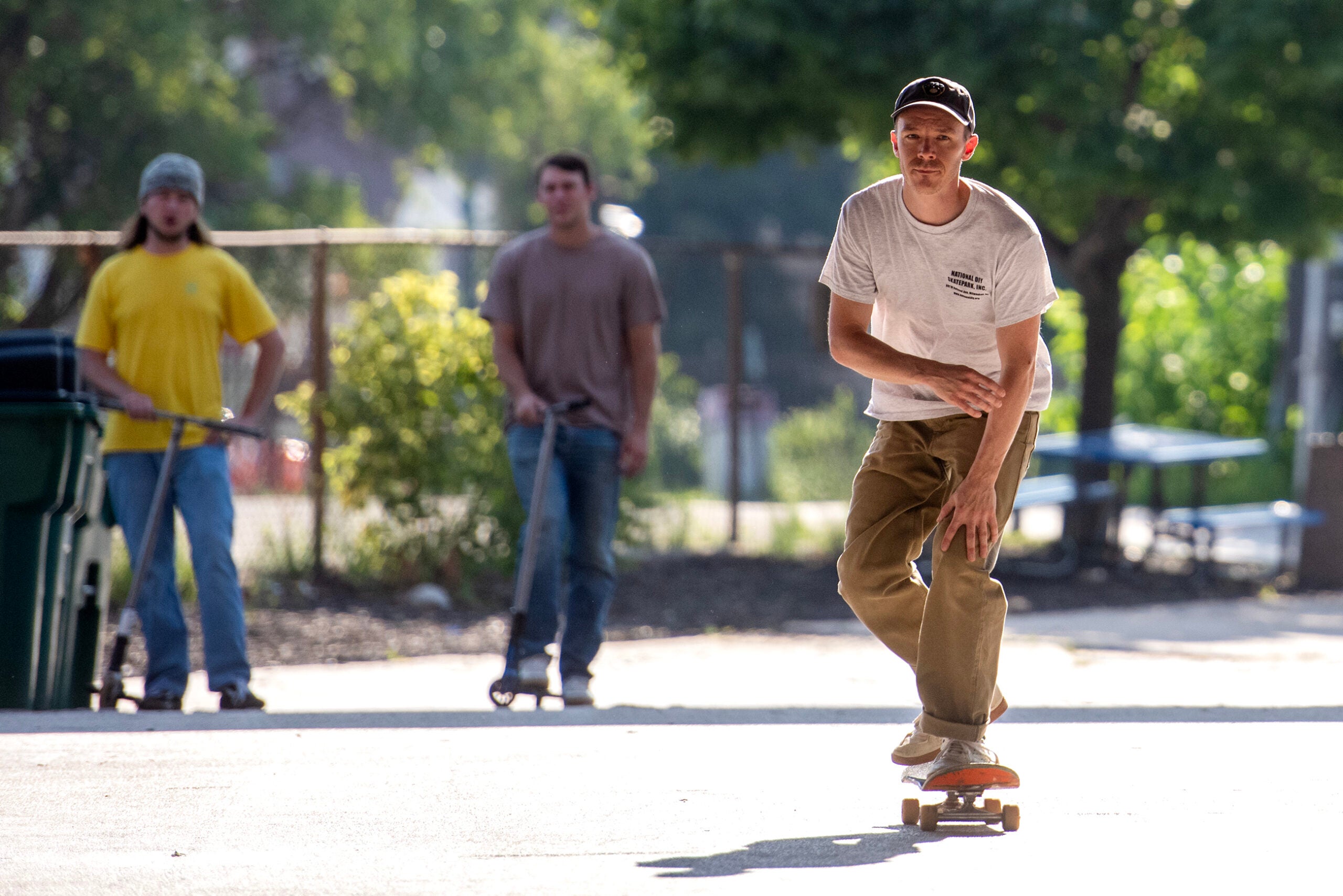 A man rides a skateboard towards the camera on a sunlit path, while two people with scooters stand in the background watching him.