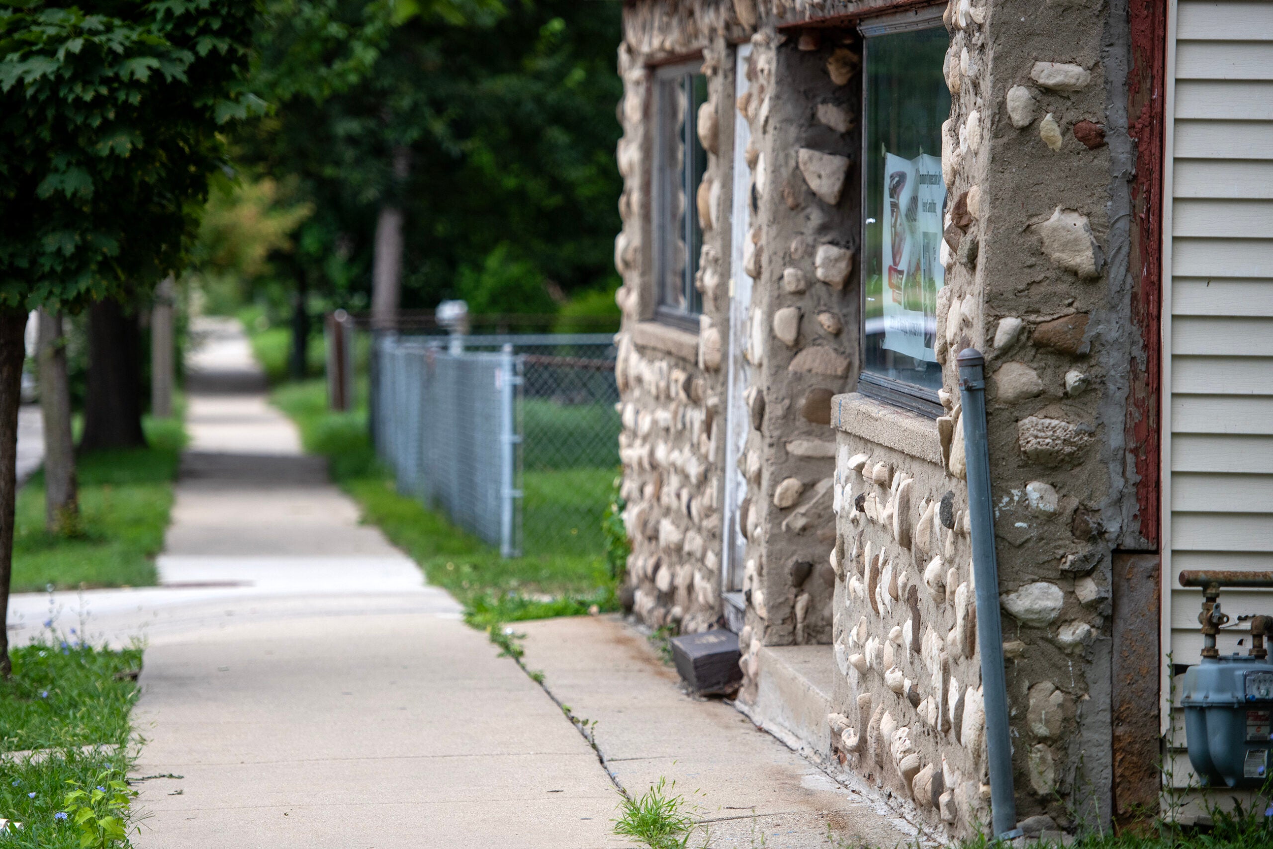 Sidewalk curving past a stone-faced building with large windows, a chain-link fence, grass, and trees in the background.