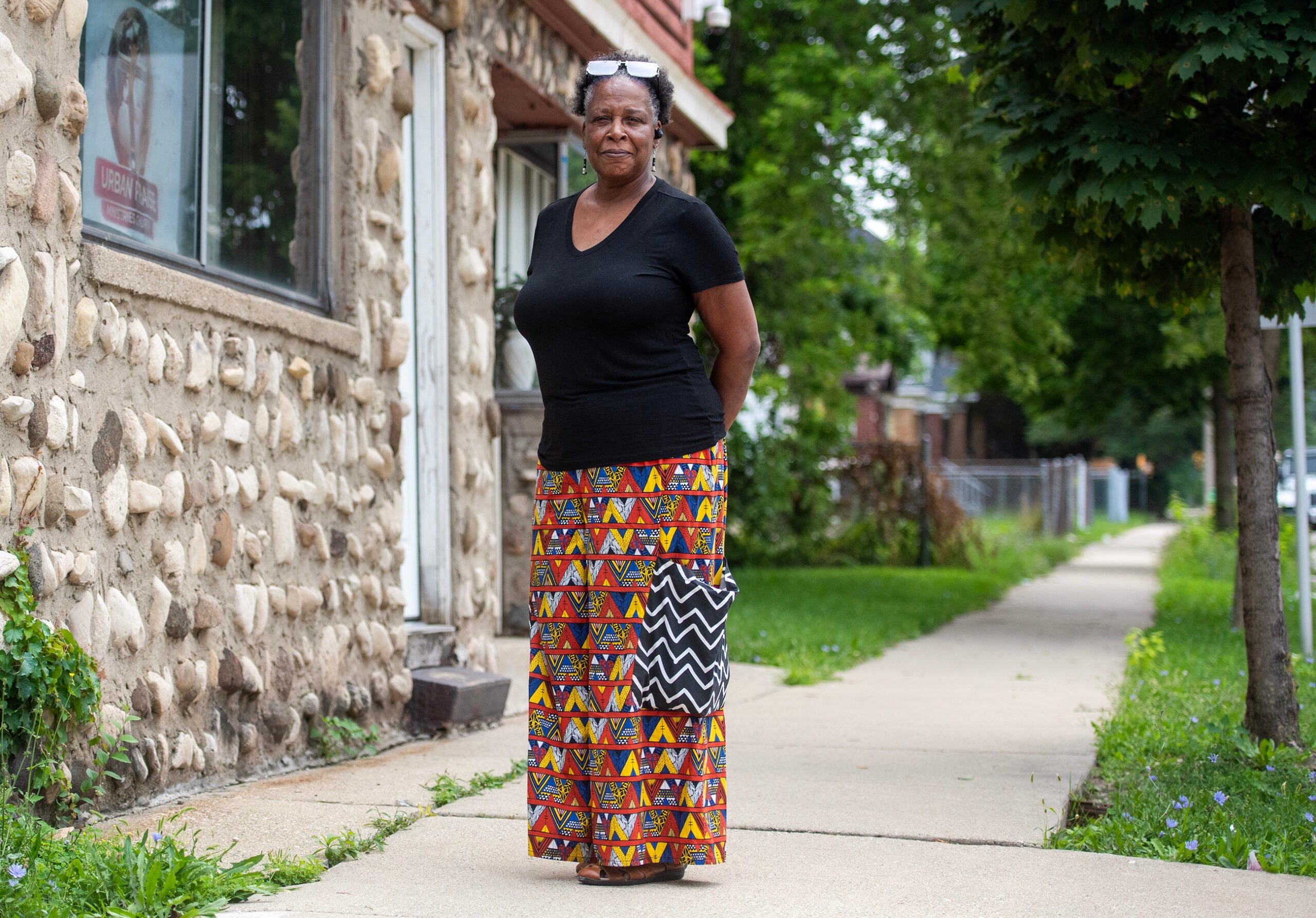 A woman stands on a sidewalk beside a stone building, wearing a black shirt and a colorful patterned skirt, with greenery and trees in the background.