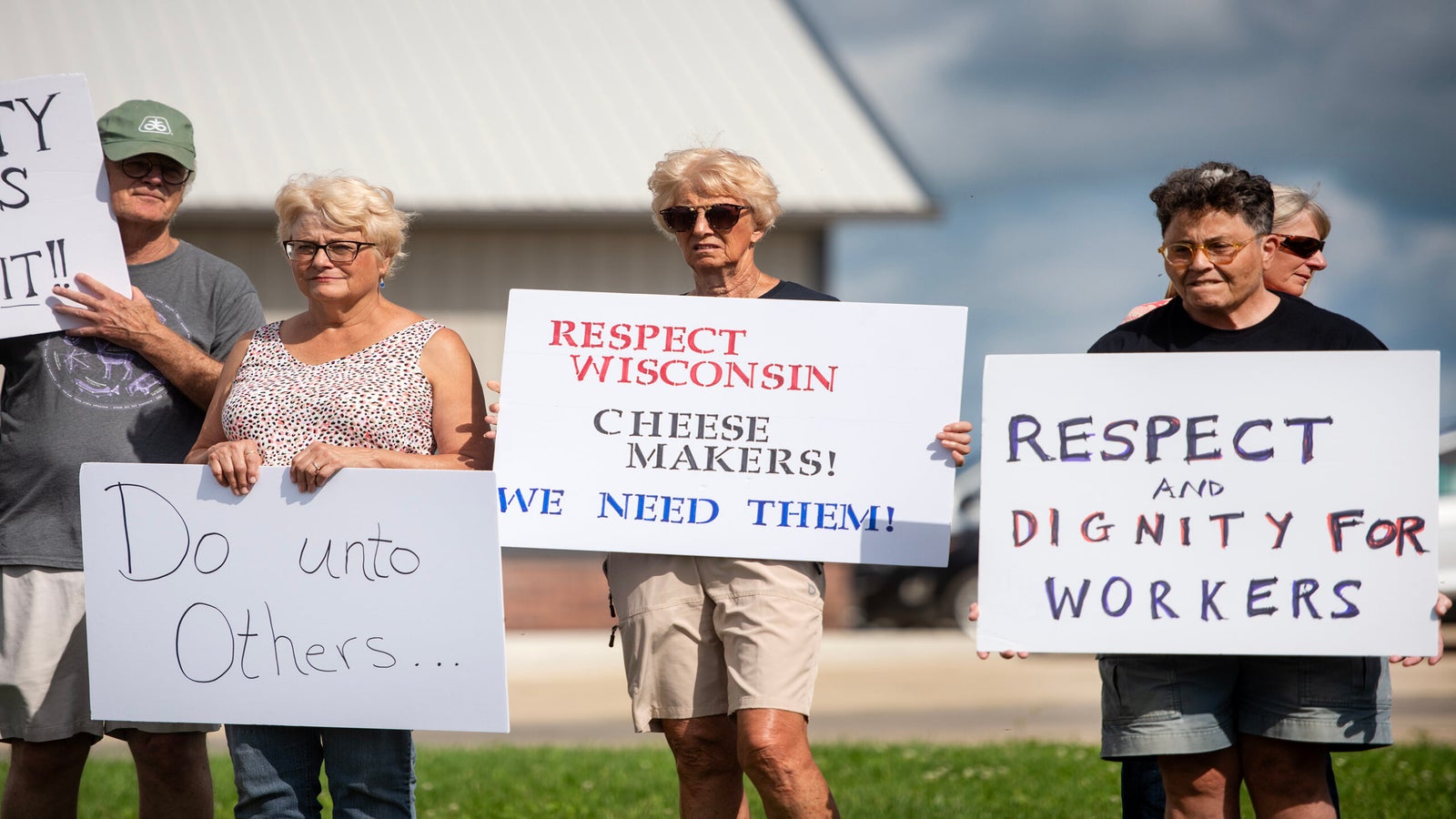 A group of people stand outside holding signs that support cheese makers and call for respect, dignity, and kindness for workers and others.