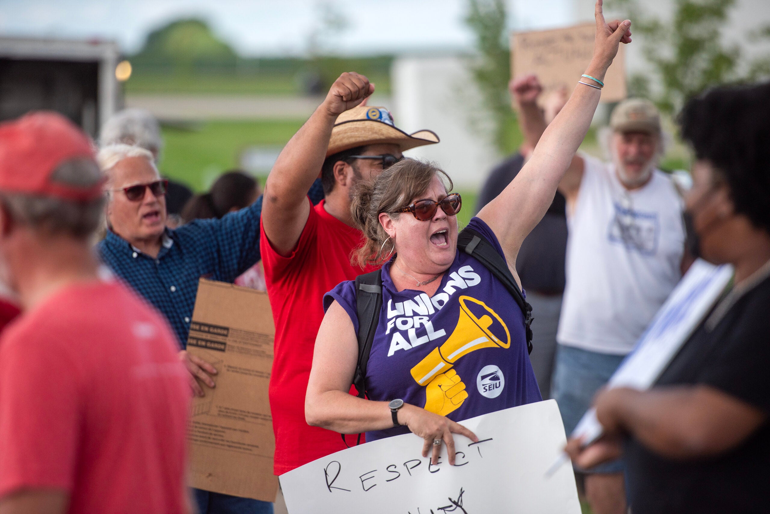 A group of people protest outdoors; a woman in front wearing a Unions For All shirt raises her arm and holds a sign that reads Respect.