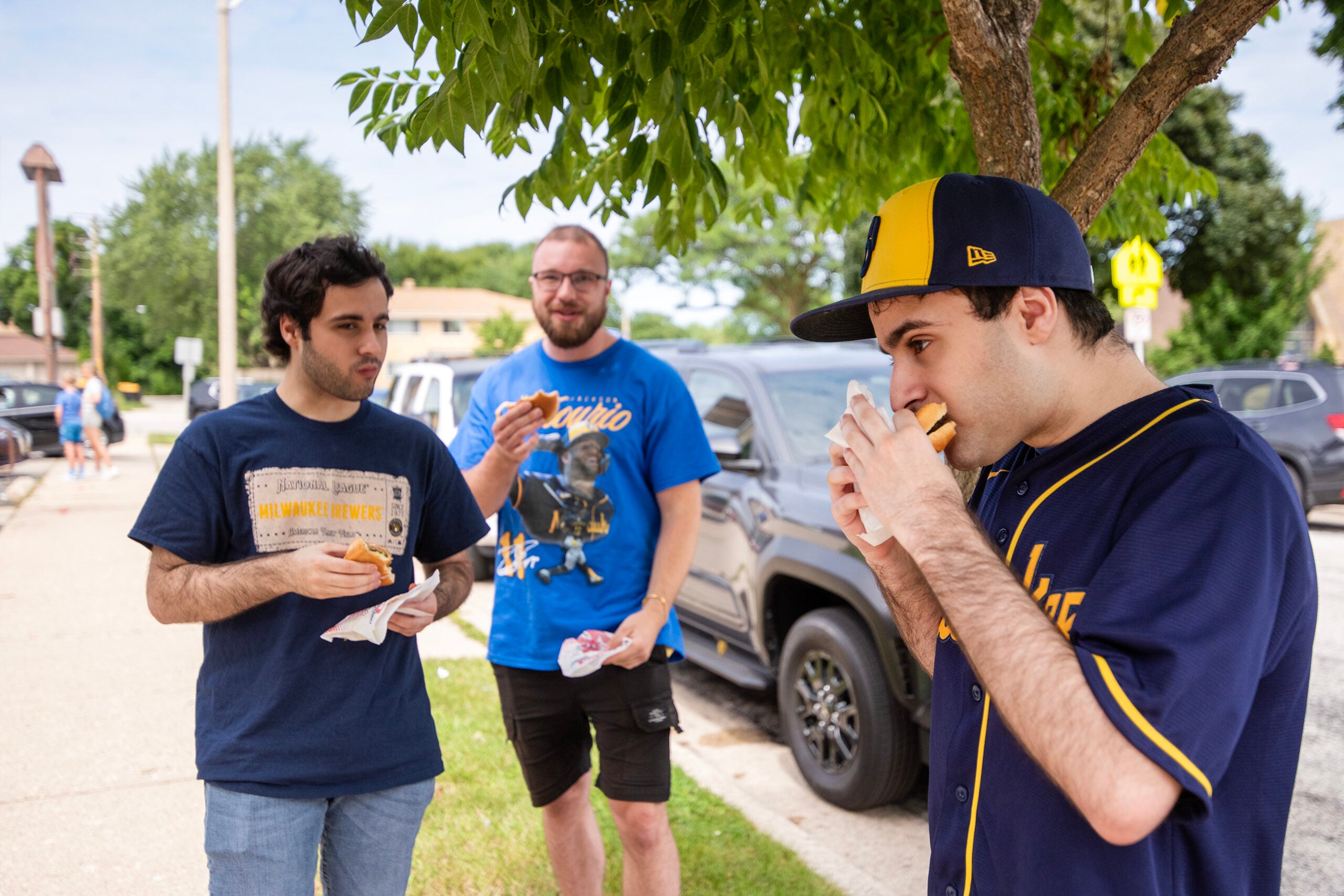 Three young men stand outside near parked cars, eating sandwiches under a leafy tree on a sunny day.