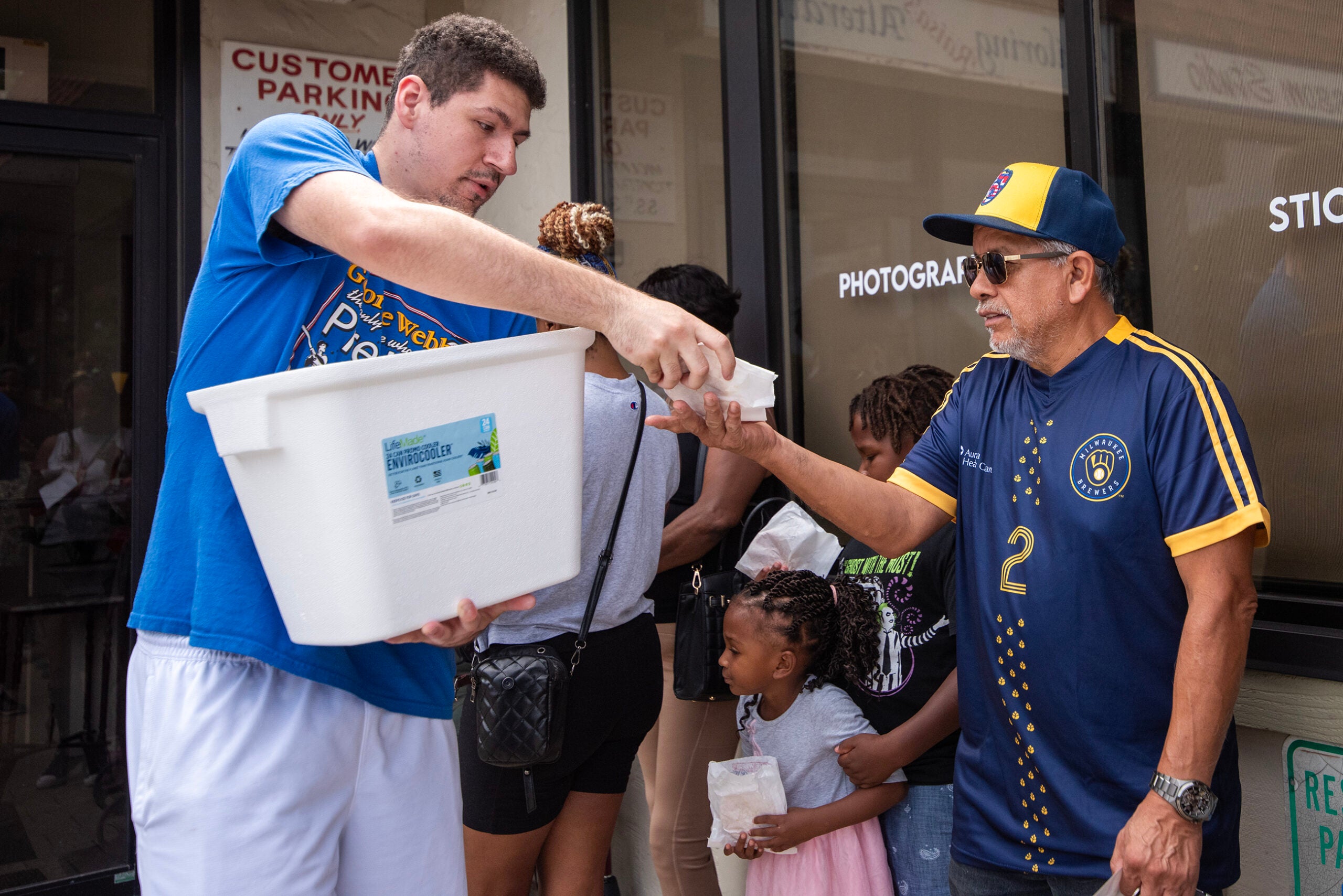 A man hands a small container to another man from a large white bin outside a building, while people including a child wait in line.