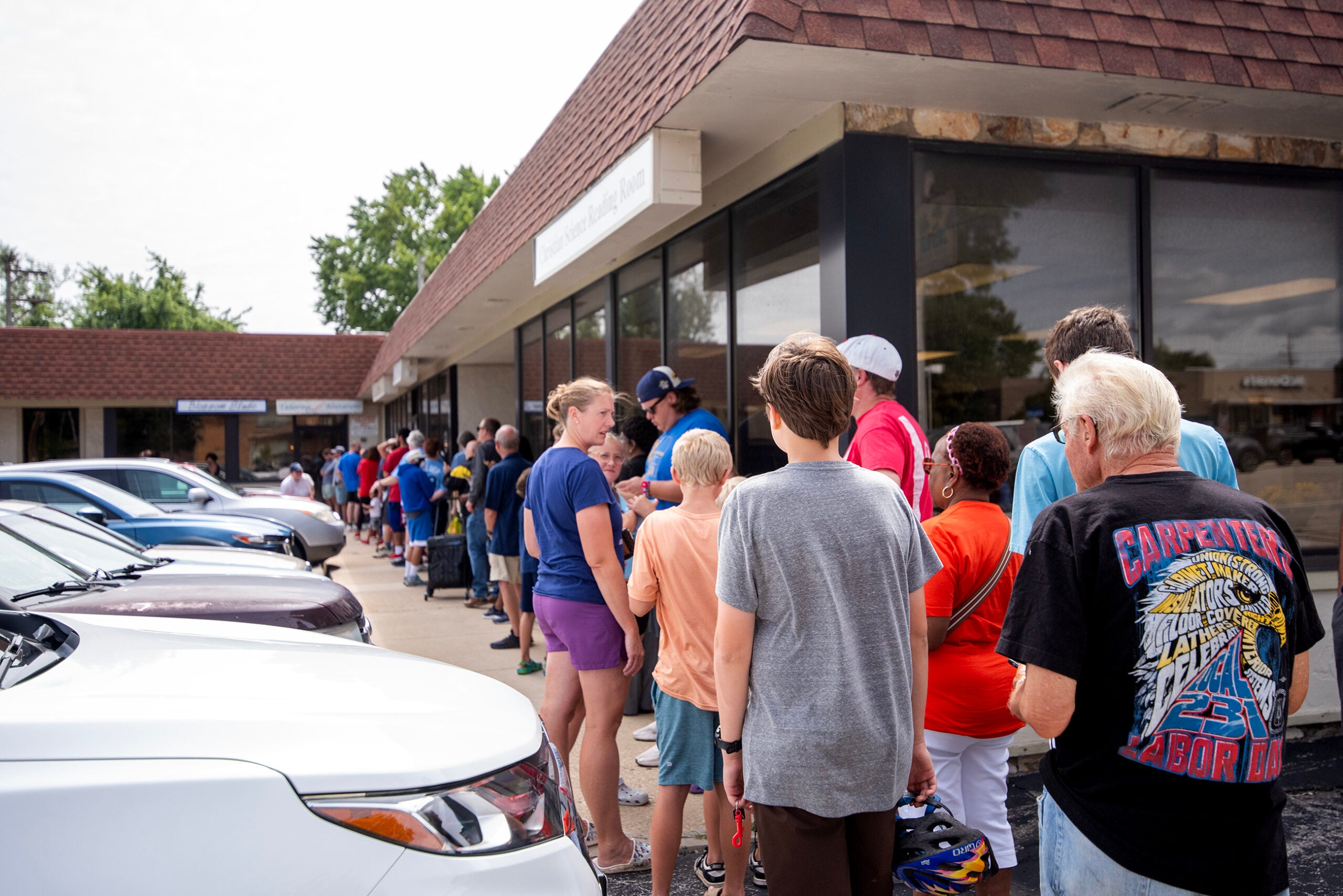 A long line of people waits outside a building on a sidewalk near parked cars on a cloudy day.