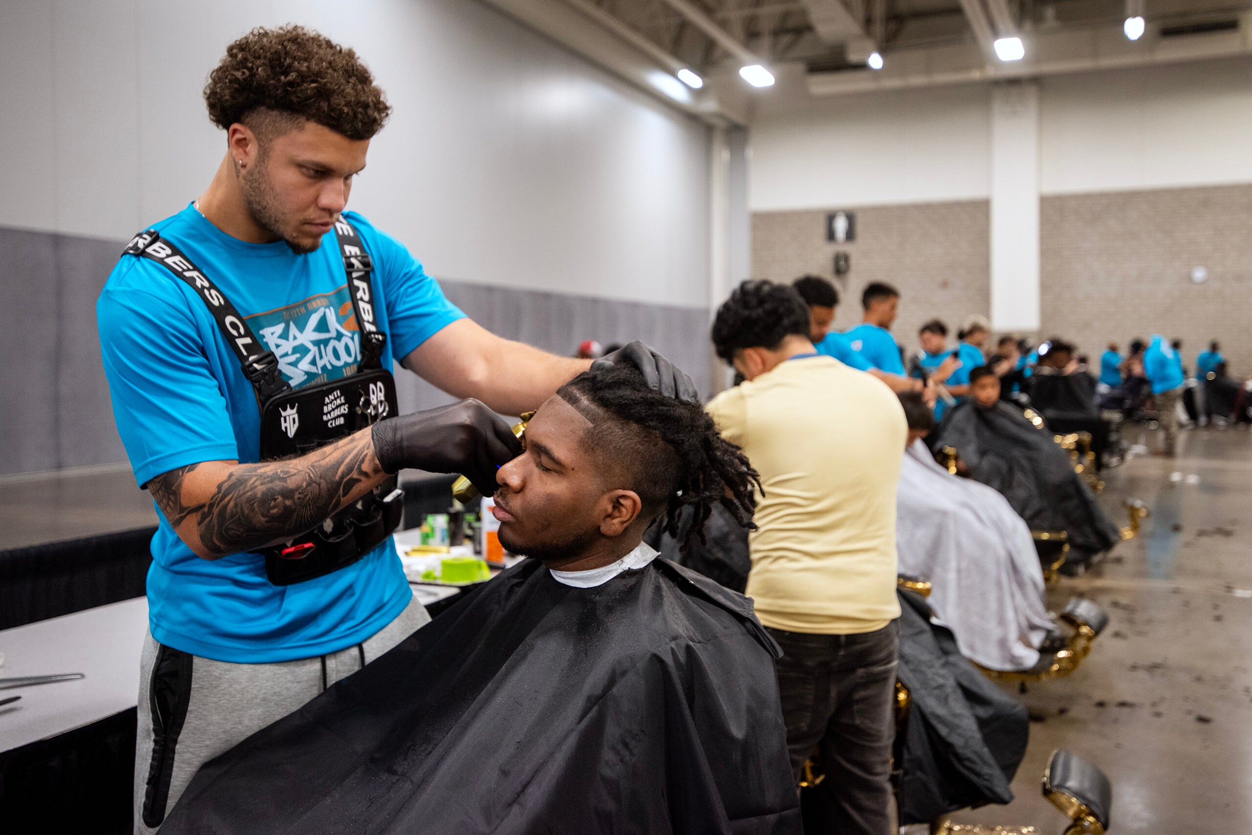 A barber trims a mans hair in a large room with several other barbers and clients in the background.