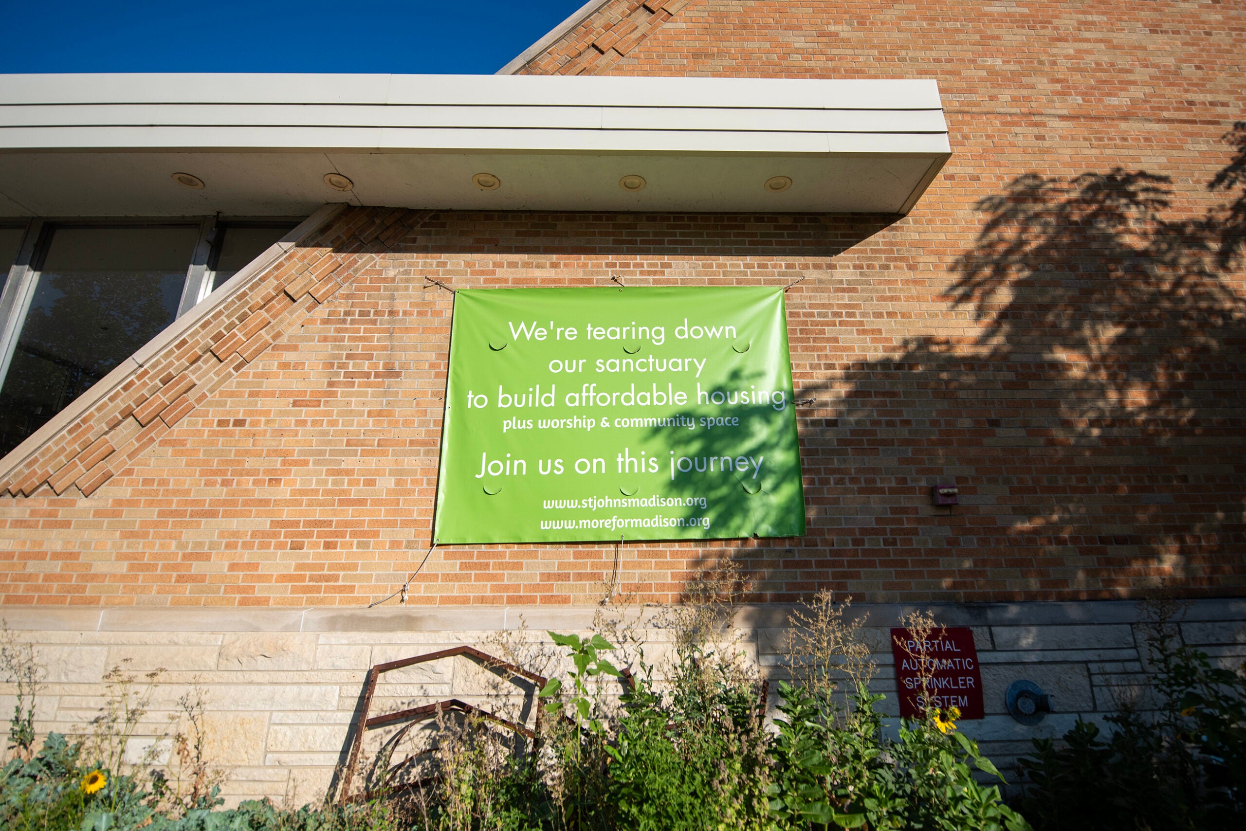 A green sign on a brick wall announces plans to tear down a sanctuary to build affordable housing, inviting people to join the project.