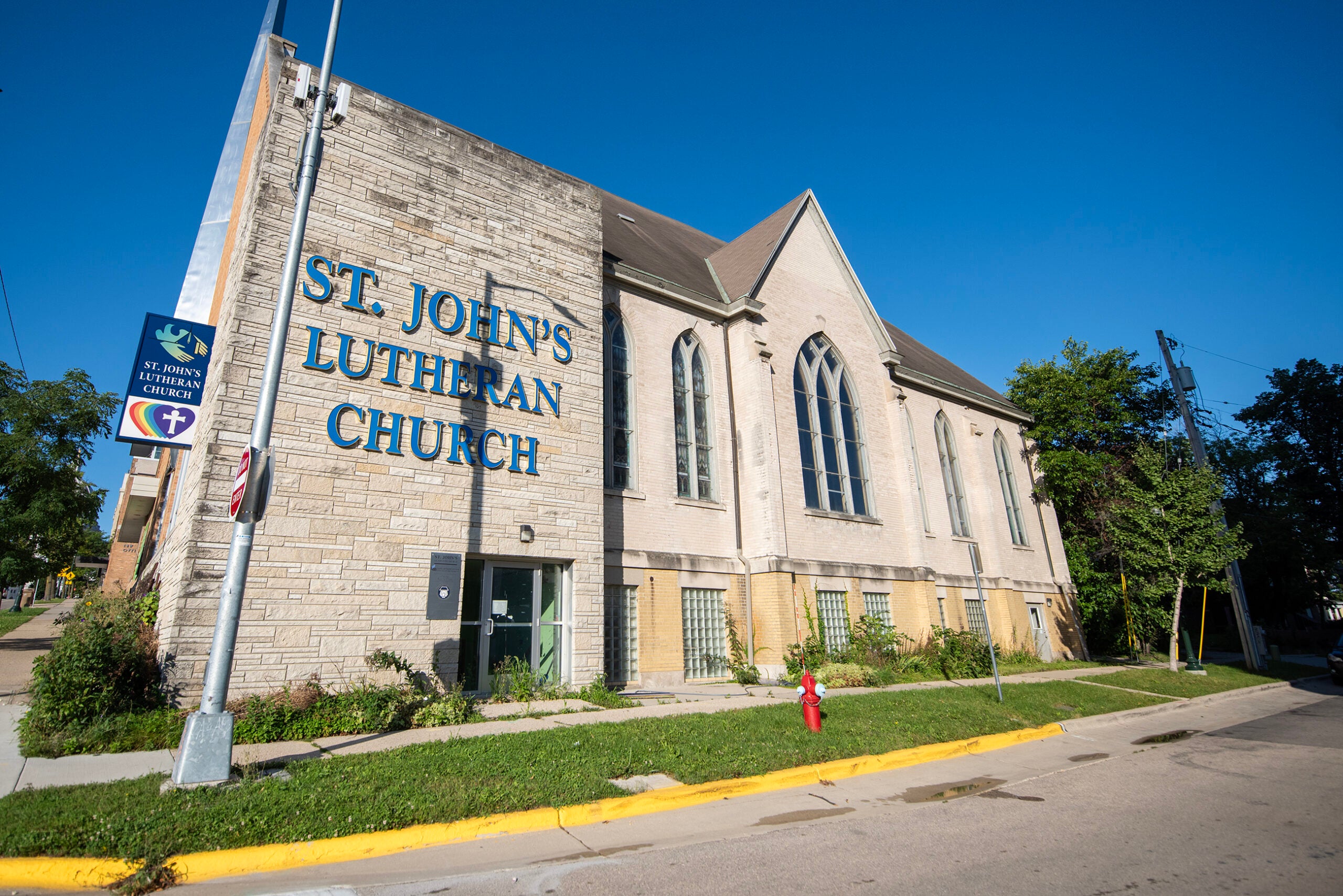A beige brick building with blue letters reading St. Johns Lutheran Church is shown on a sunny day, with a church sign and a fire hydrant in front.