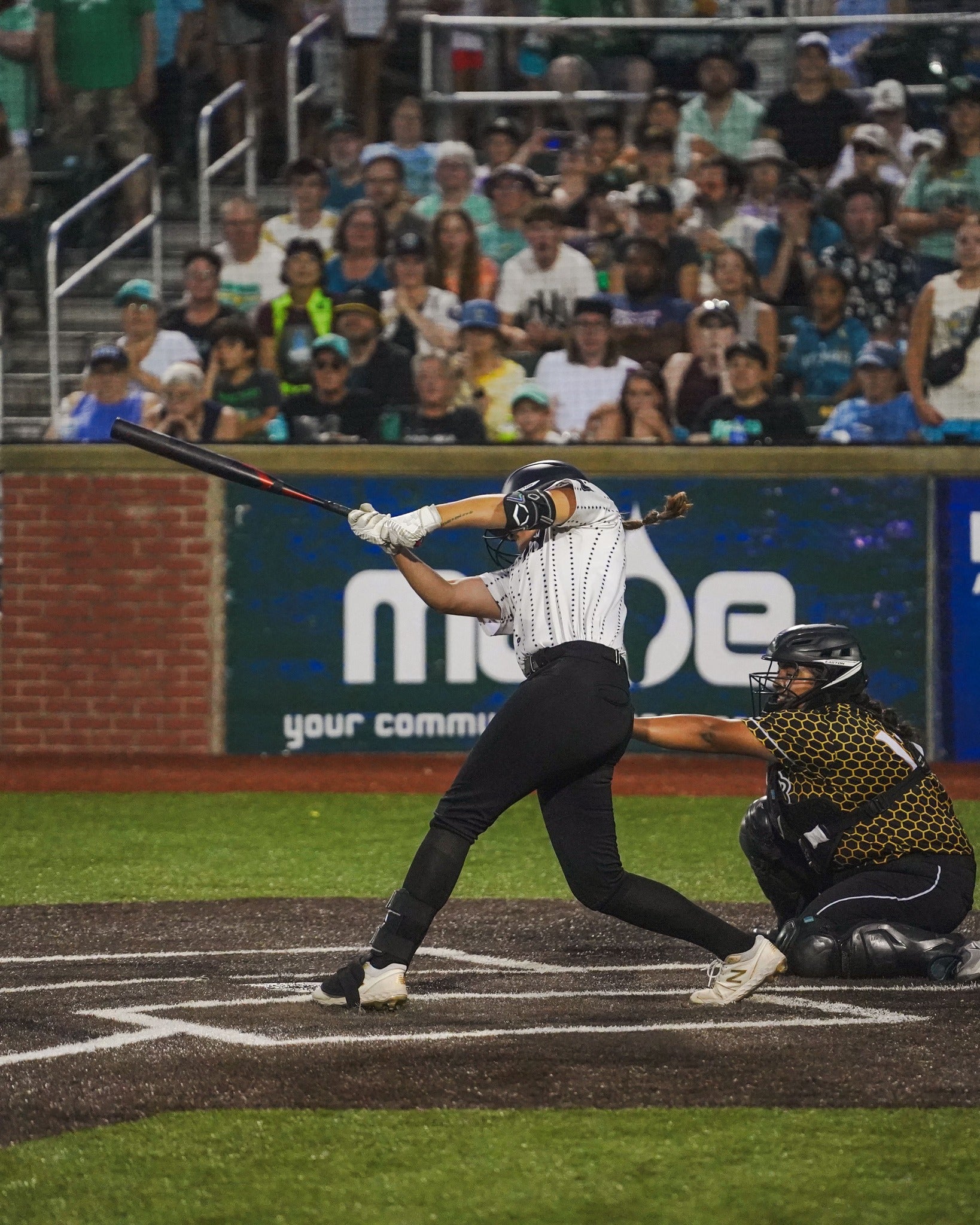 A baseball player swings a bat at home plate while the catcher crouches behind, with a crowd watching from the stands.