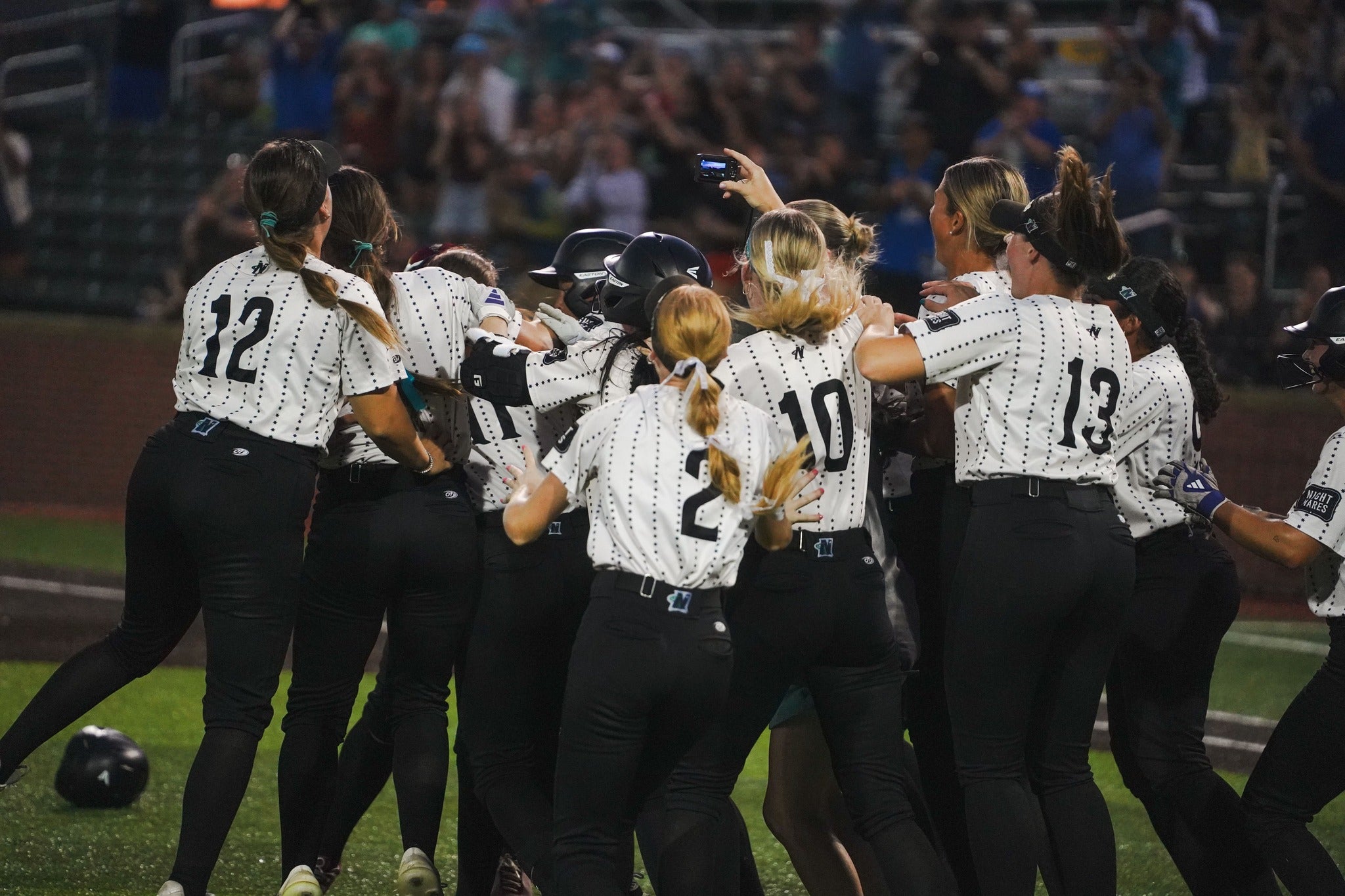 A softball team in black and white uniforms celebrates together on the field, with players hugging and cheering after a game.