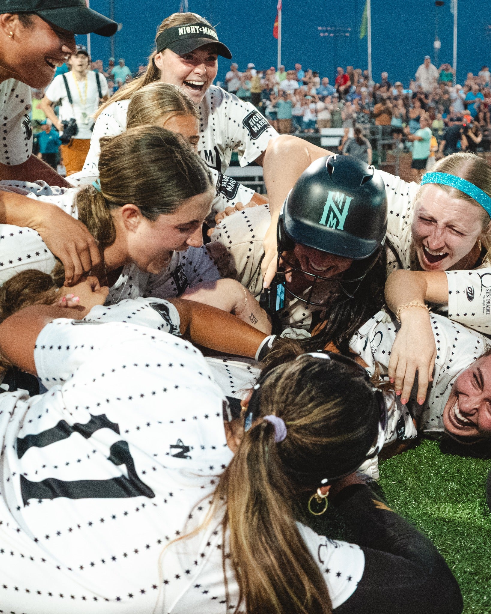 A group of softball players in white uniforms pile on top of each other and celebrate on the field, with a crowd visible in the background.