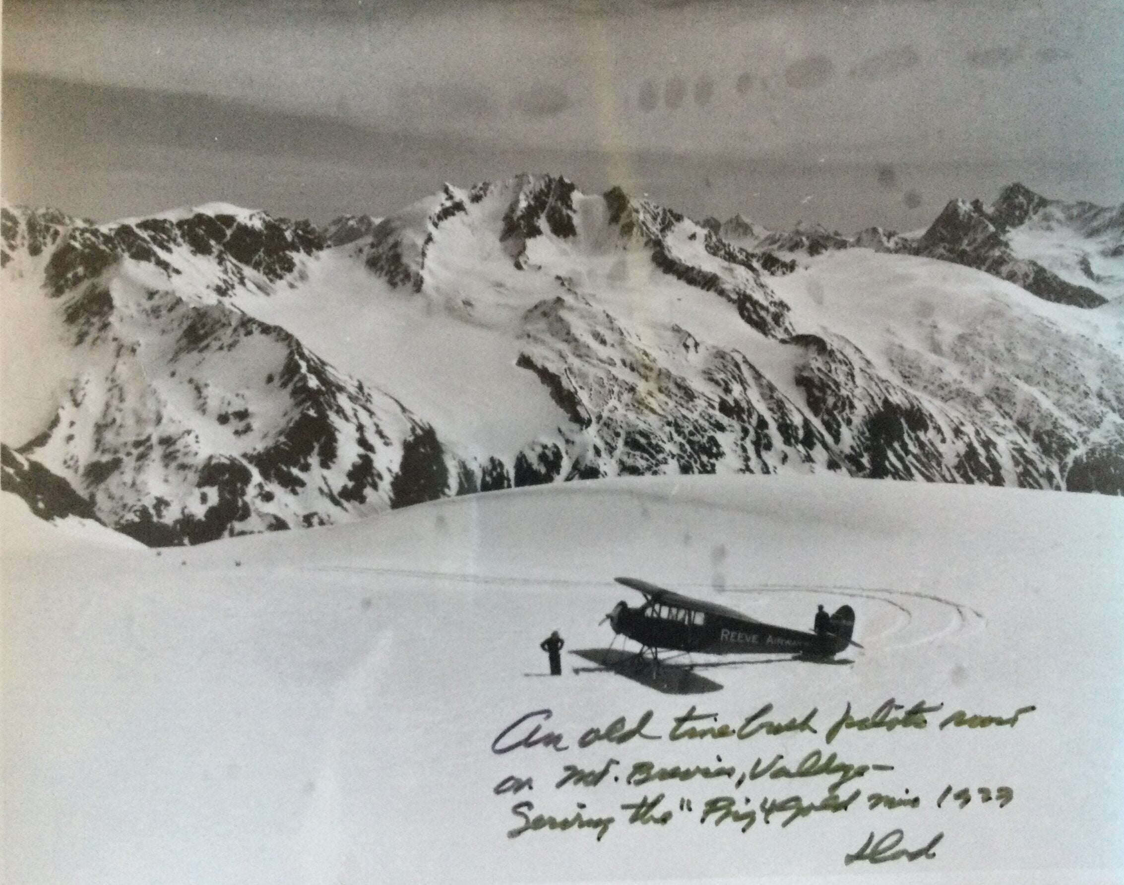 A small vintage twin-engine plane is parked on a snowy mountain plateau with rugged peaks in the background; handwritten notes are visible at the bottom of the photo.