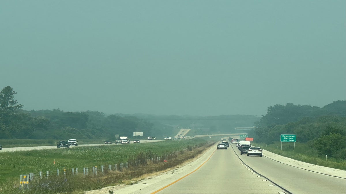 Cars drive on a divided highway surrounded by greenery under a hazy sky, with visibility reduced due to air pollution or haze.