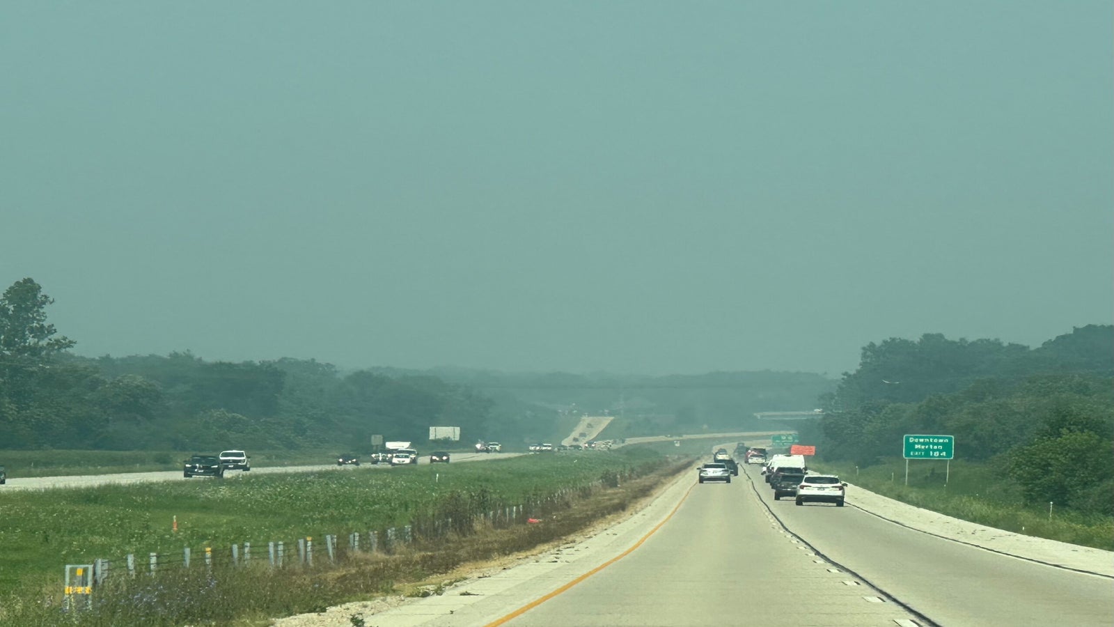 Cars drive on a divided highway surrounded by greenery under a hazy sky, with visibility reduced due to air pollution or haze.