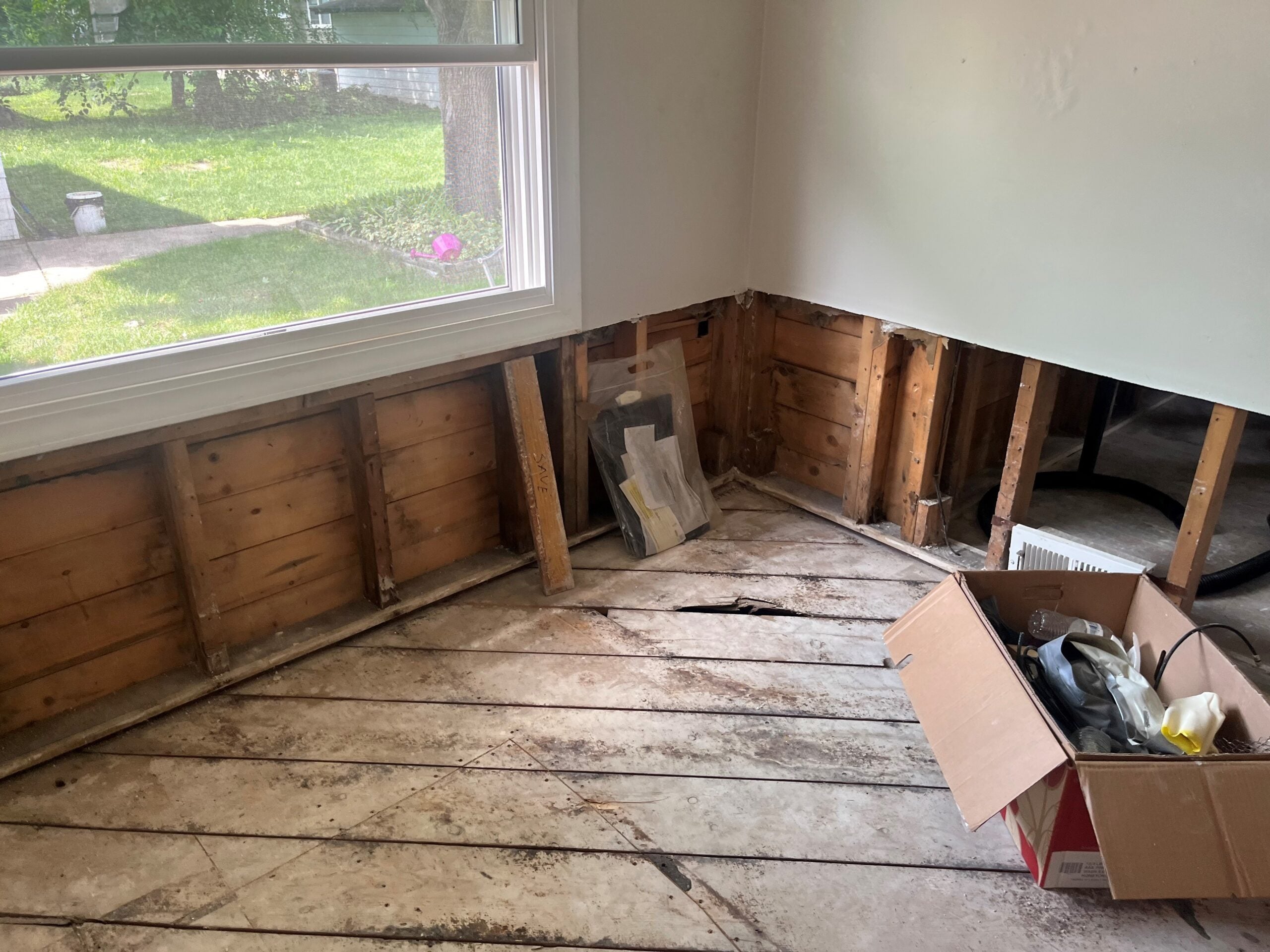 Partially renovated room with exposed wall studs and floorboards, a large window, and a cardboard box containing various tools and materials.