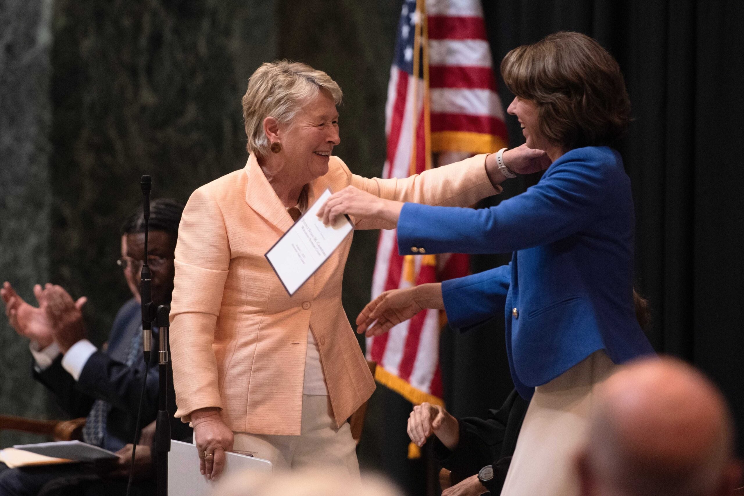 Two women smile and move to embrace on stage, each holding papers, with an American flag and applauding people in the background.