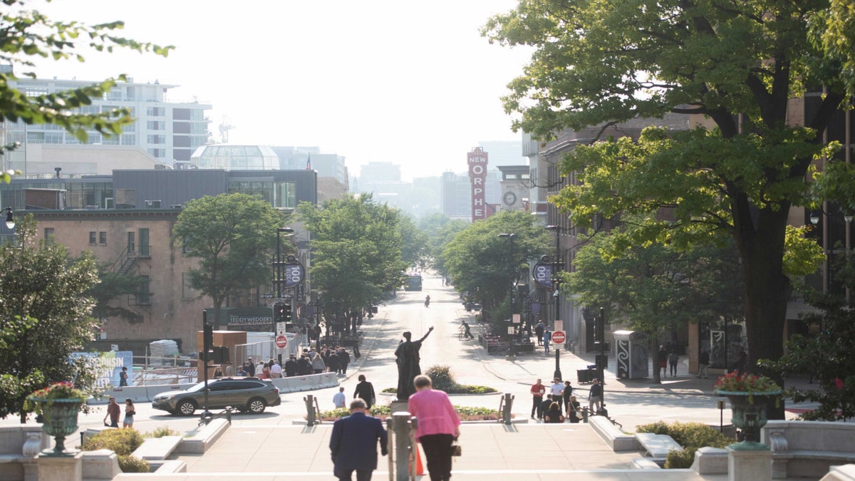 A city street scene with people walking, trees lining the road, and a statue in the center background; buildings and a theater sign are visible in the distance.