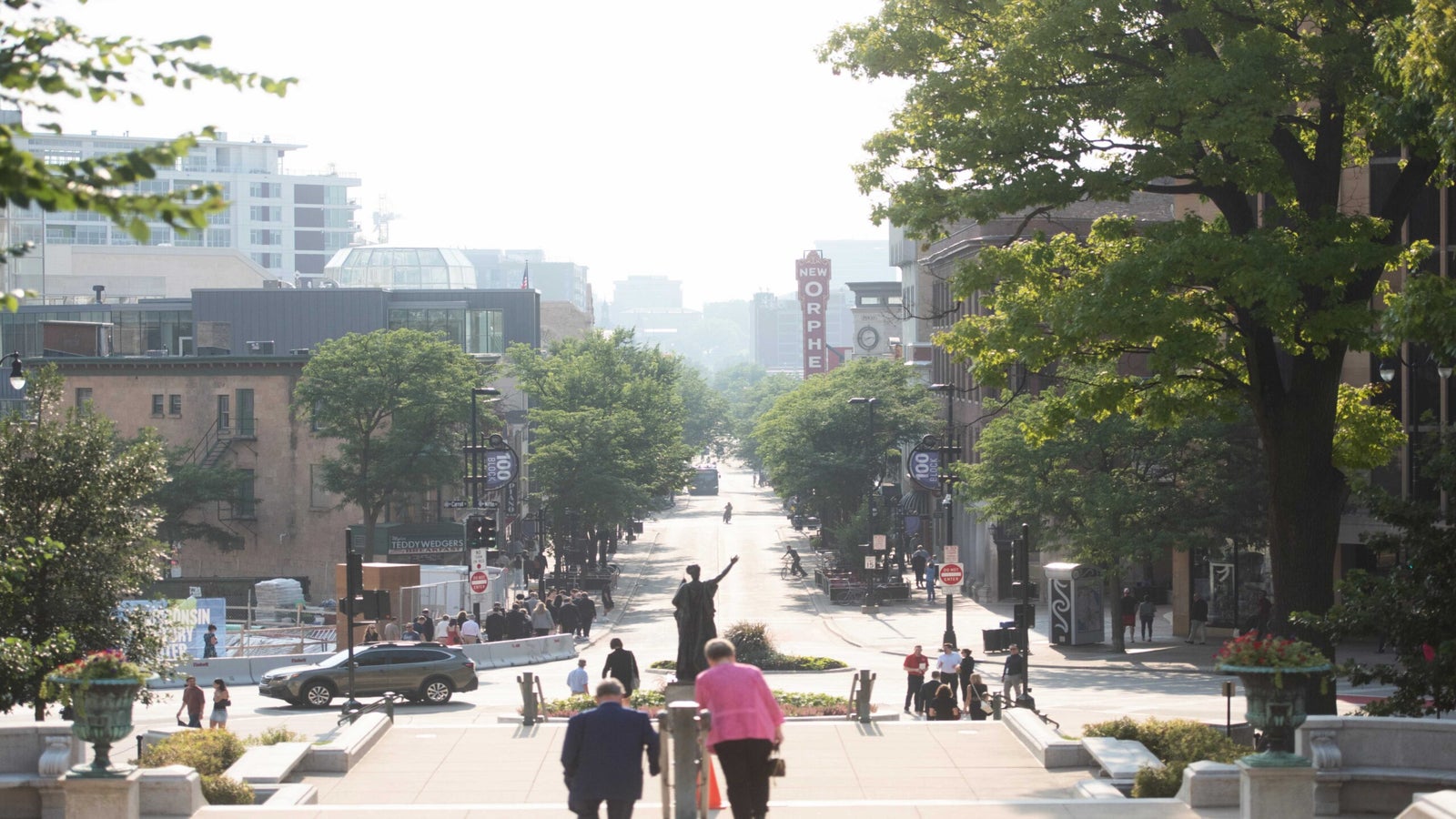 A city street scene with people walking, trees lining the road, and a statue in the center background; buildings and a theater sign are visible in the distance.